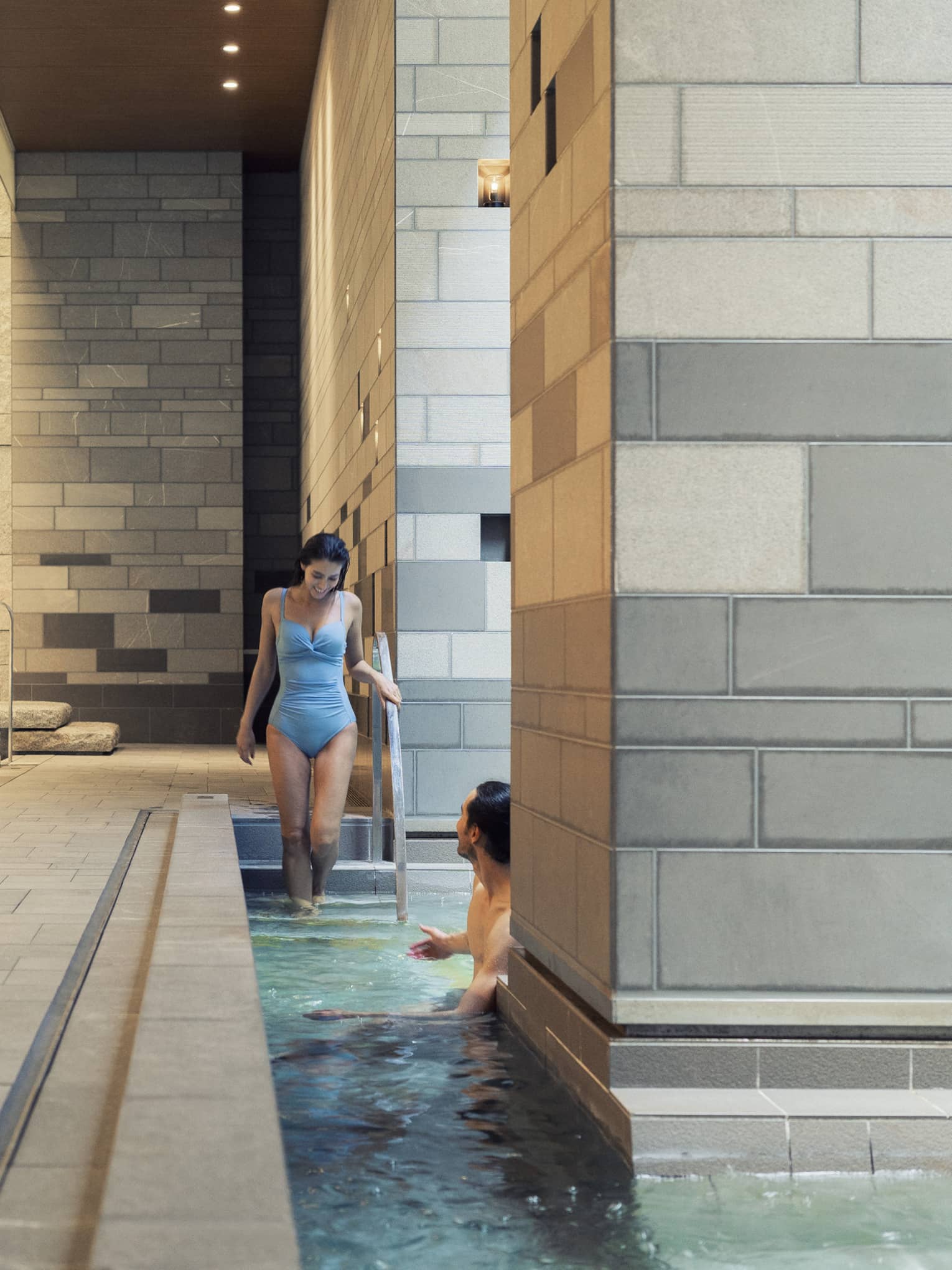 A guest enters a pool via the stairs to meet another guest who is half-hidden by a large grey-tiled column in the foreground.