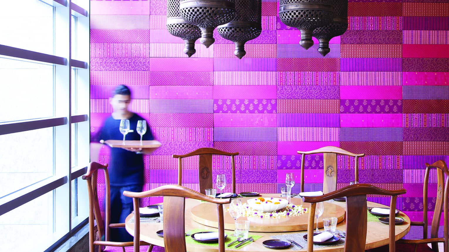 Waiter holds tray with wine glasses by large round dining table in front of purple textile wall