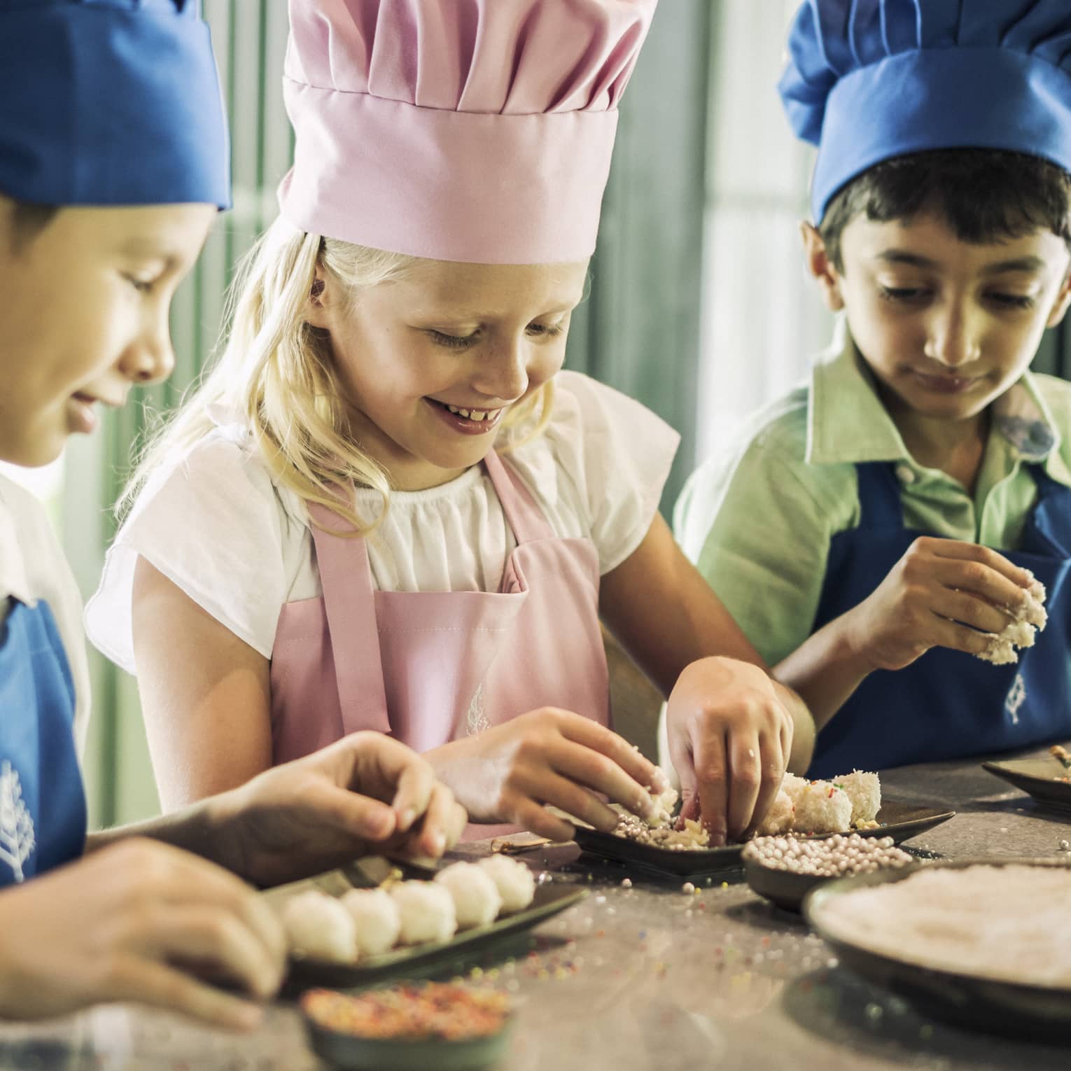 Three children in blue and pink aprons and chef hats roll pastries on small black trays
