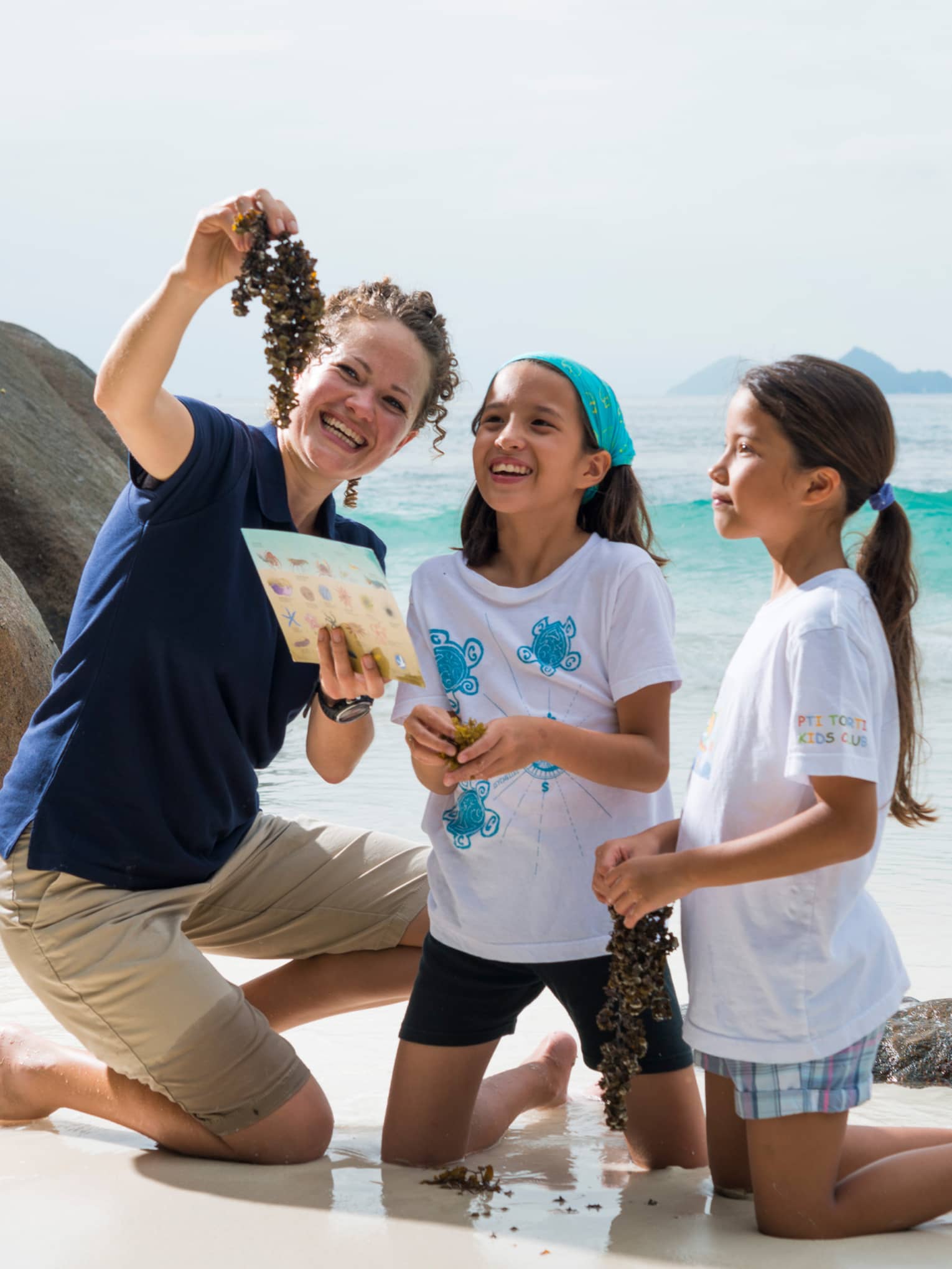 Woman kneels on white sand beach beside boulder, holds up seaweed to show two girls
