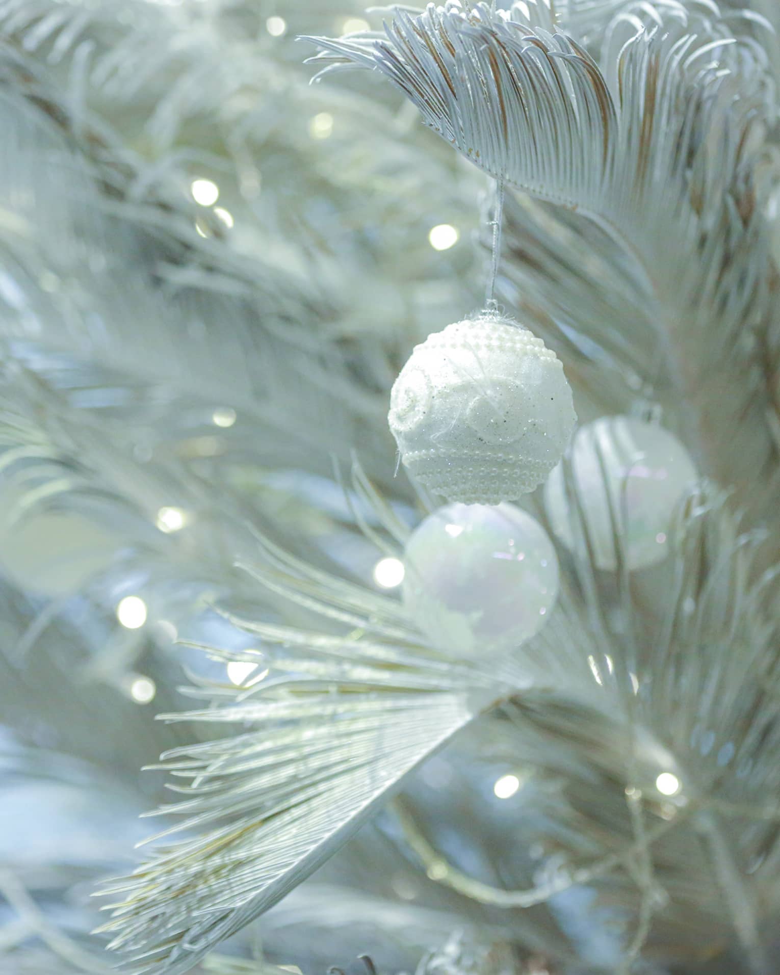 Close up of white Christmas tree branches with tiny twinkling lights and several round, white ornaments