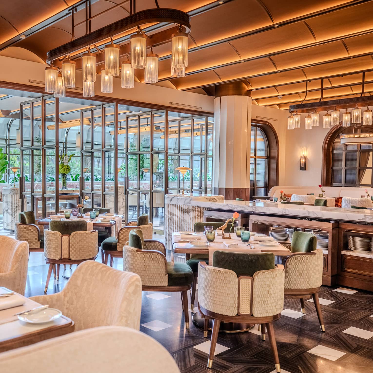 Restaurant dining room featuring tall beamed ceilings, white marble tables, dark flooring and green-and-white velvet chairs all lit by warm overhead lights