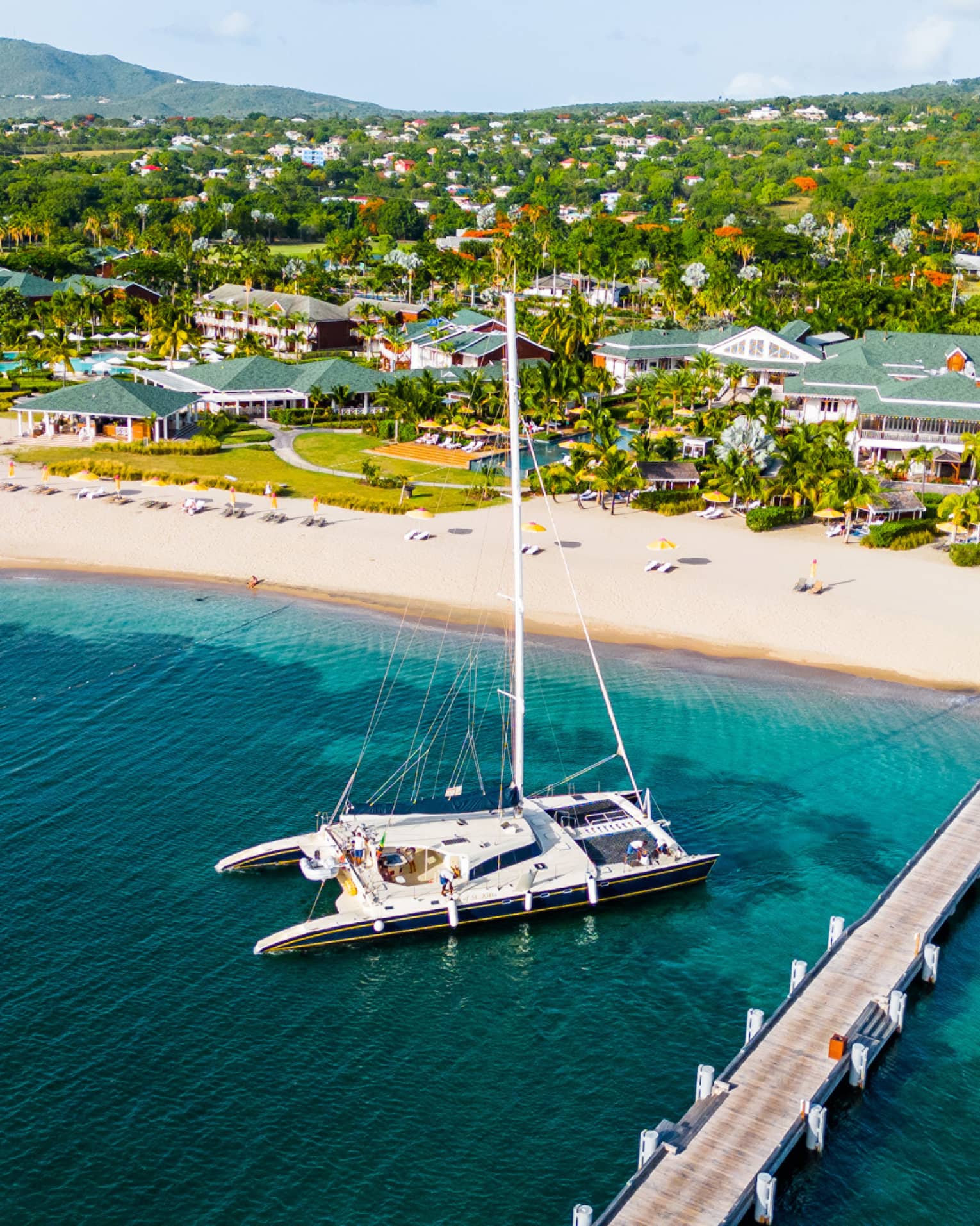 Aerial view of catamaran anchored near a dock reaching out from a beach with a sprawling resort along the coastline set among the trees