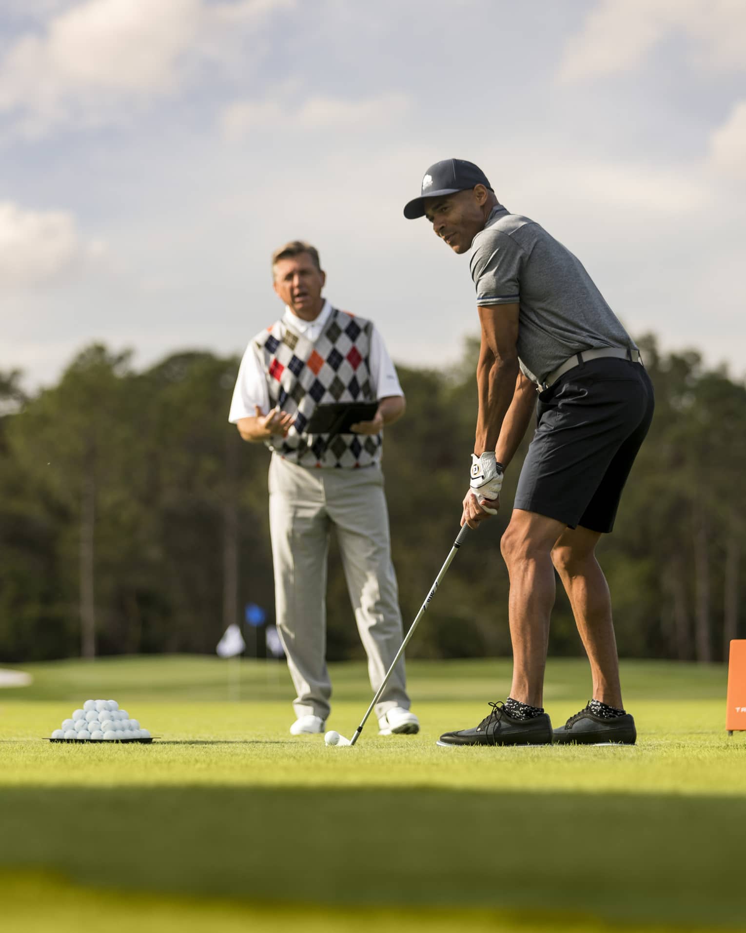 Against a clear sky, a golfer tees up, a tray of balls and a stand of clubs nearby. An instructor stands in the background.