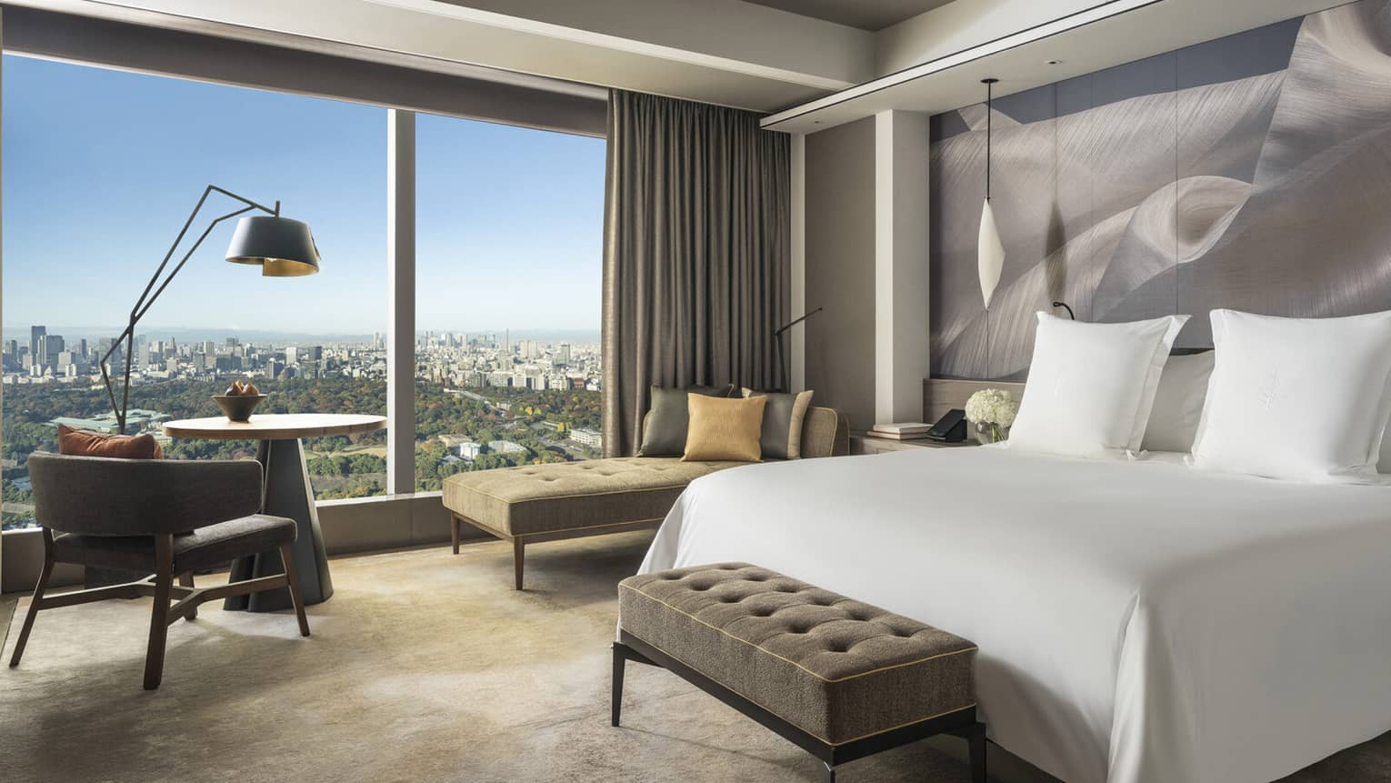 Guest room with king bed, tufted bench, chaise lounge, table with armchair, pendant lights and large window with city views