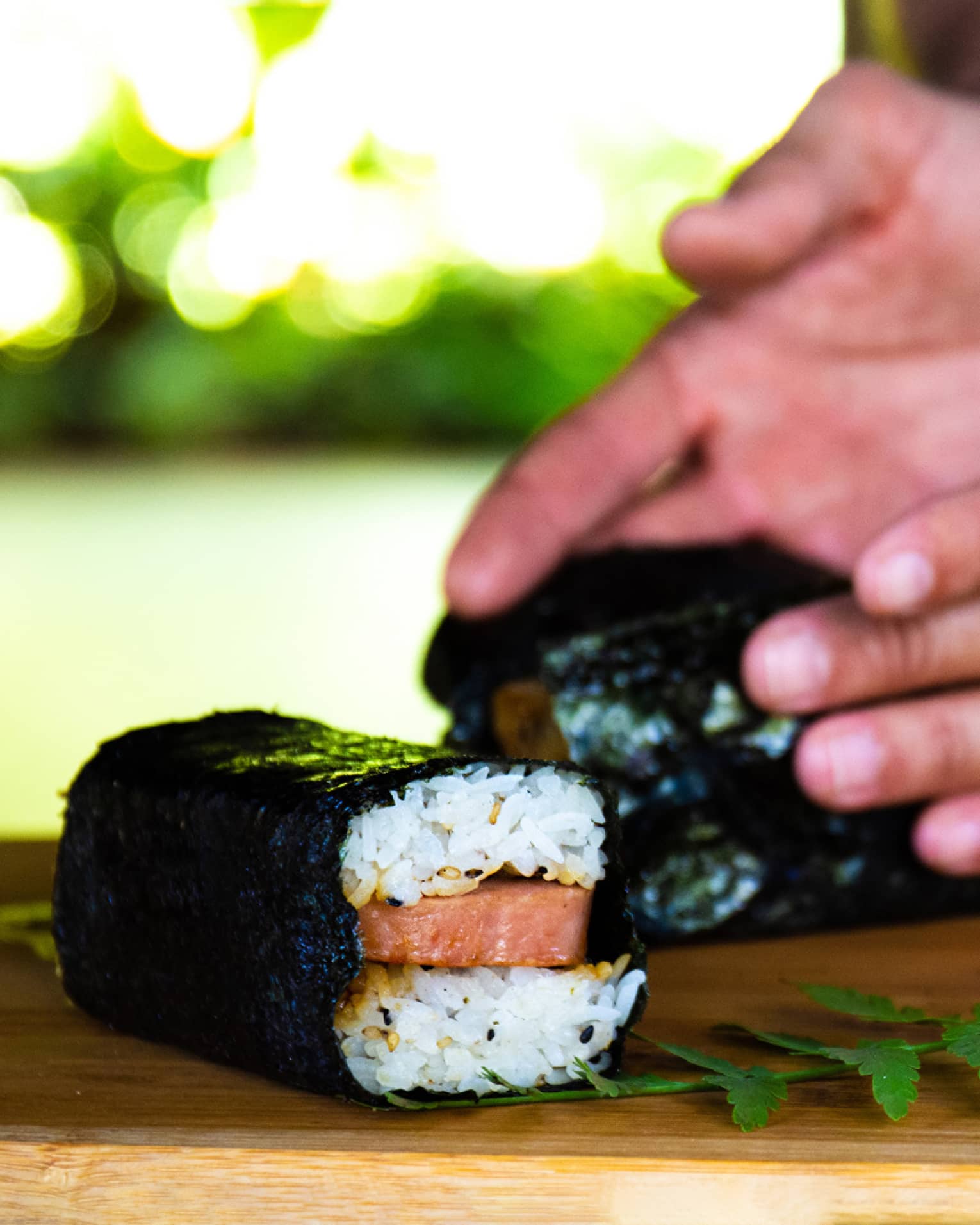 Close-up of hands wrapping nori around layered rice sandwiching raw salmon, beside a green fern frond on a wood board.