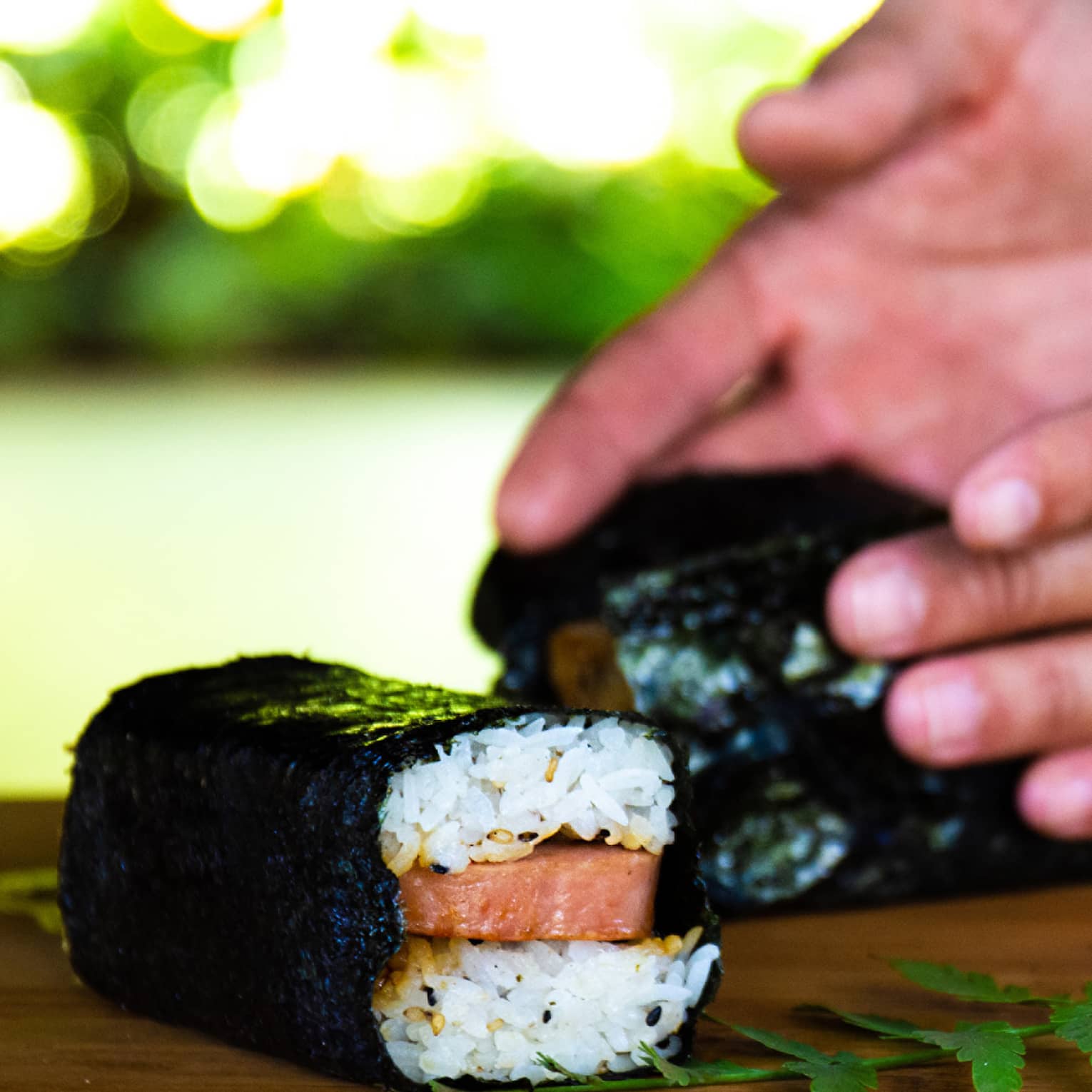 Close-up of hands wrapping nori around layered rice sandwiching raw salmon, beside a green fern frond on a wood board.