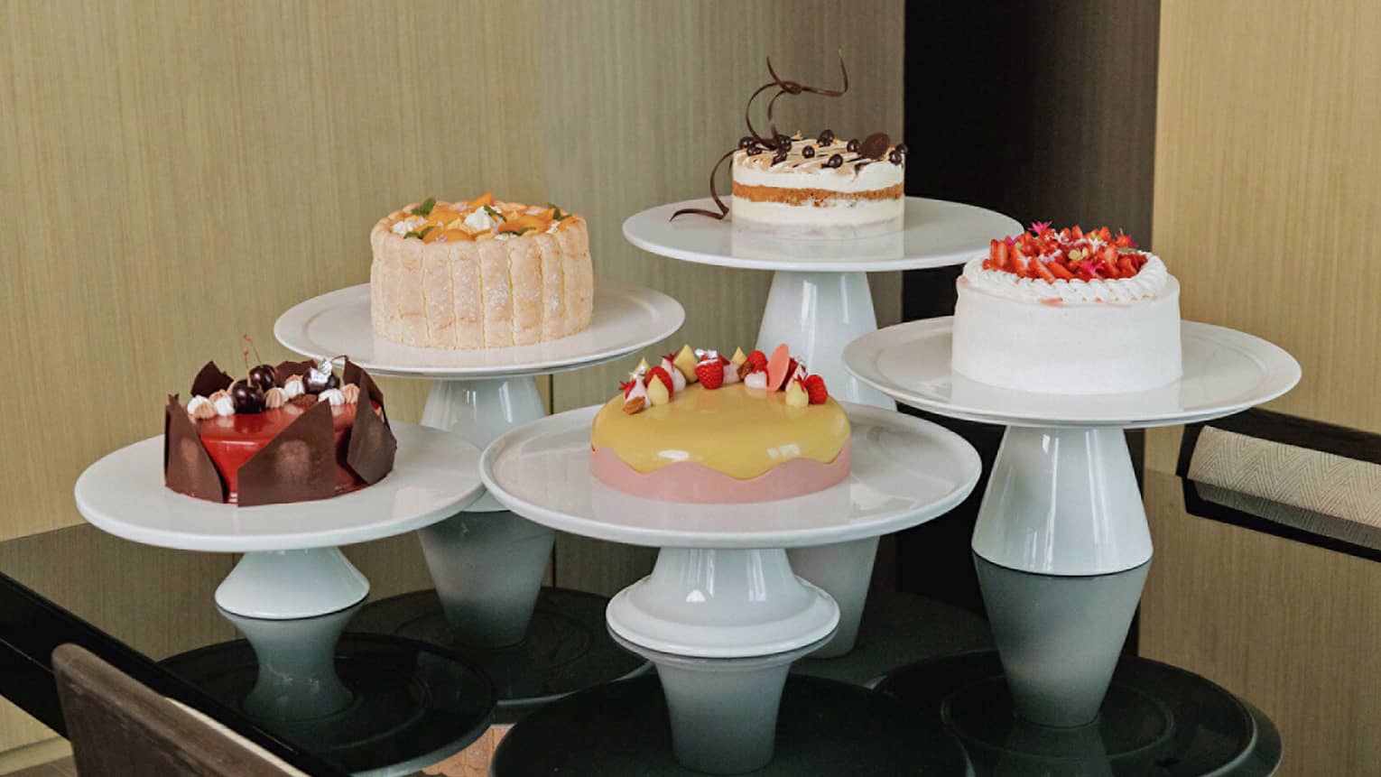 Five cakes of various colors and flavours, each on its own round, white cake stand are arranged together in a group on a black table