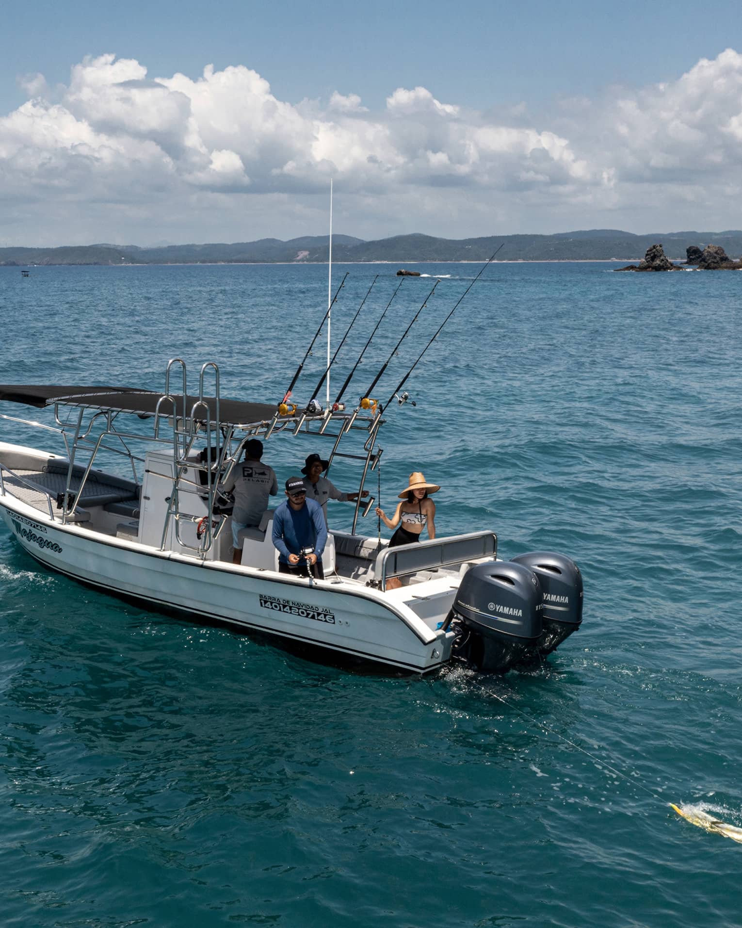 Guest reels in a large yellow fish from the back of a boat in open water near rocky islands, while two friends look on.