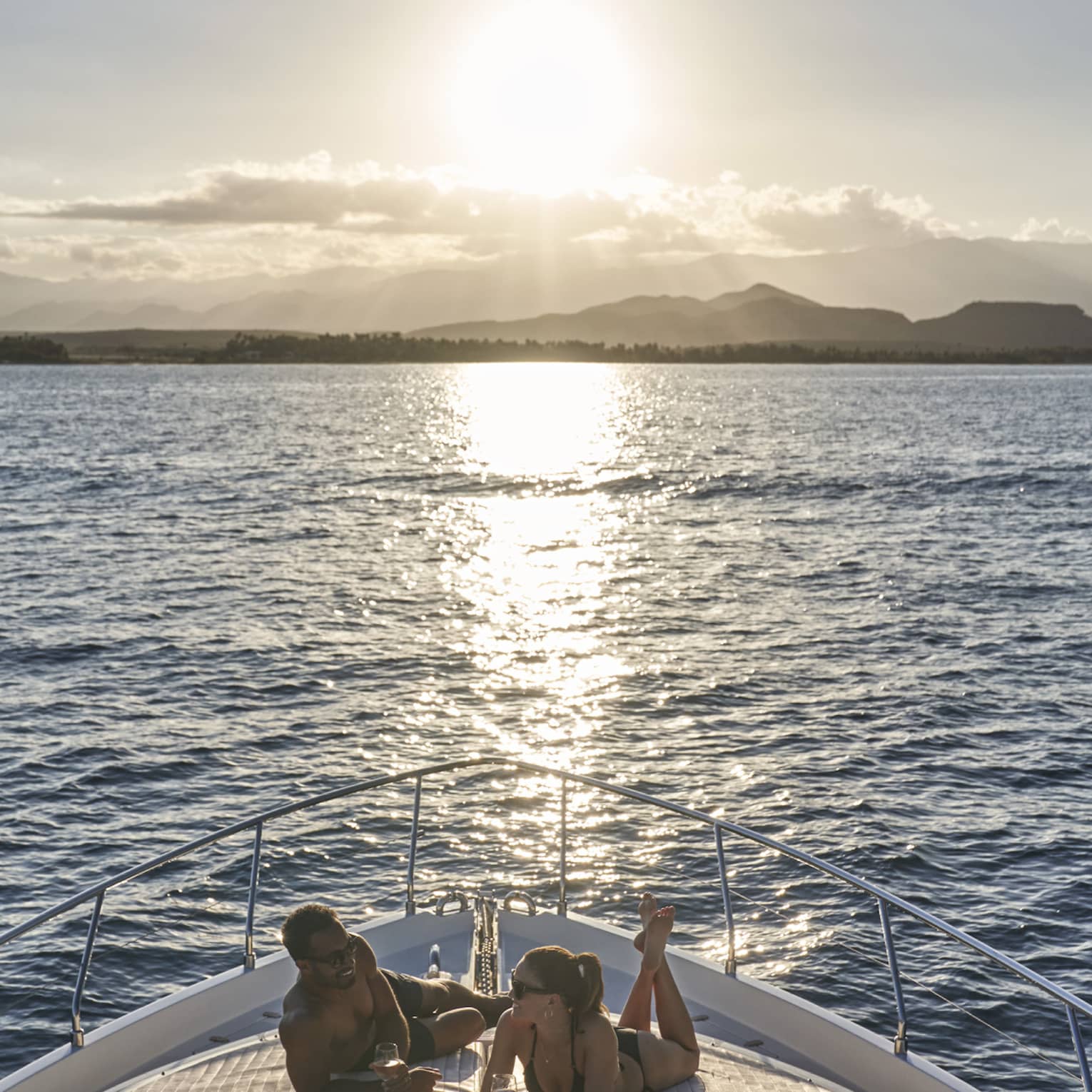 Couple wearing swimsuits relaxes on front yacht deck as sun sets over ocean