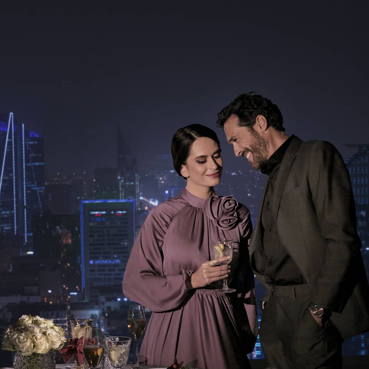 A beautiful view of the cityscape at night, a loving couple stands in front of the view with a romantically set dinner table.