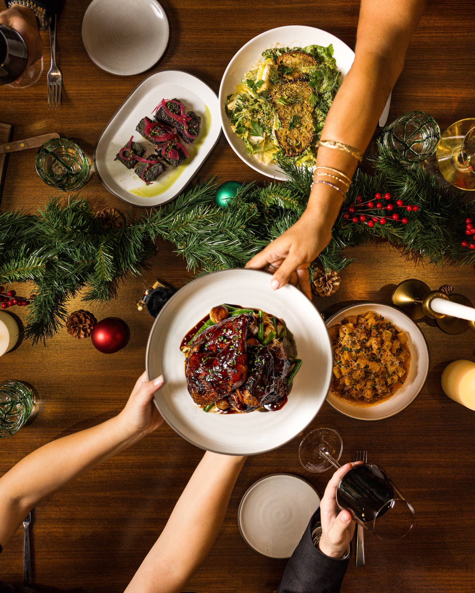 Hands pass a dish of food to another over a table set for a holiday meal with a green garland in the centre