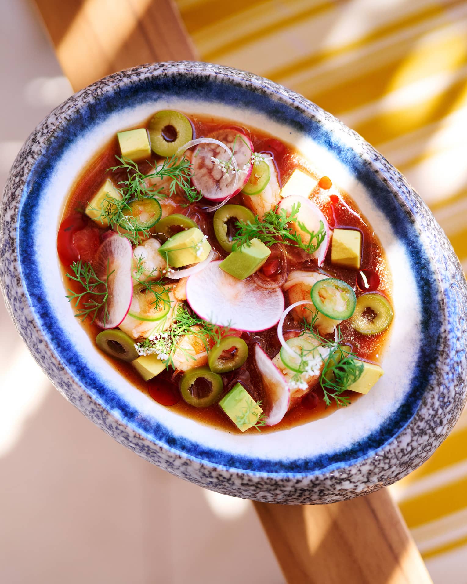 Top-down view of red pozole soup in an elegant ceramic bowl and garnished with radish, avocado, jalapeños and green olives.