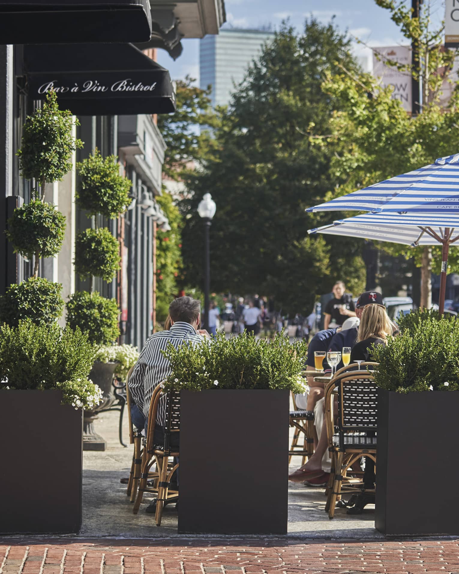 People dine on Bar a Vin Bistrot sidewalk patio behind planters, umbrellas