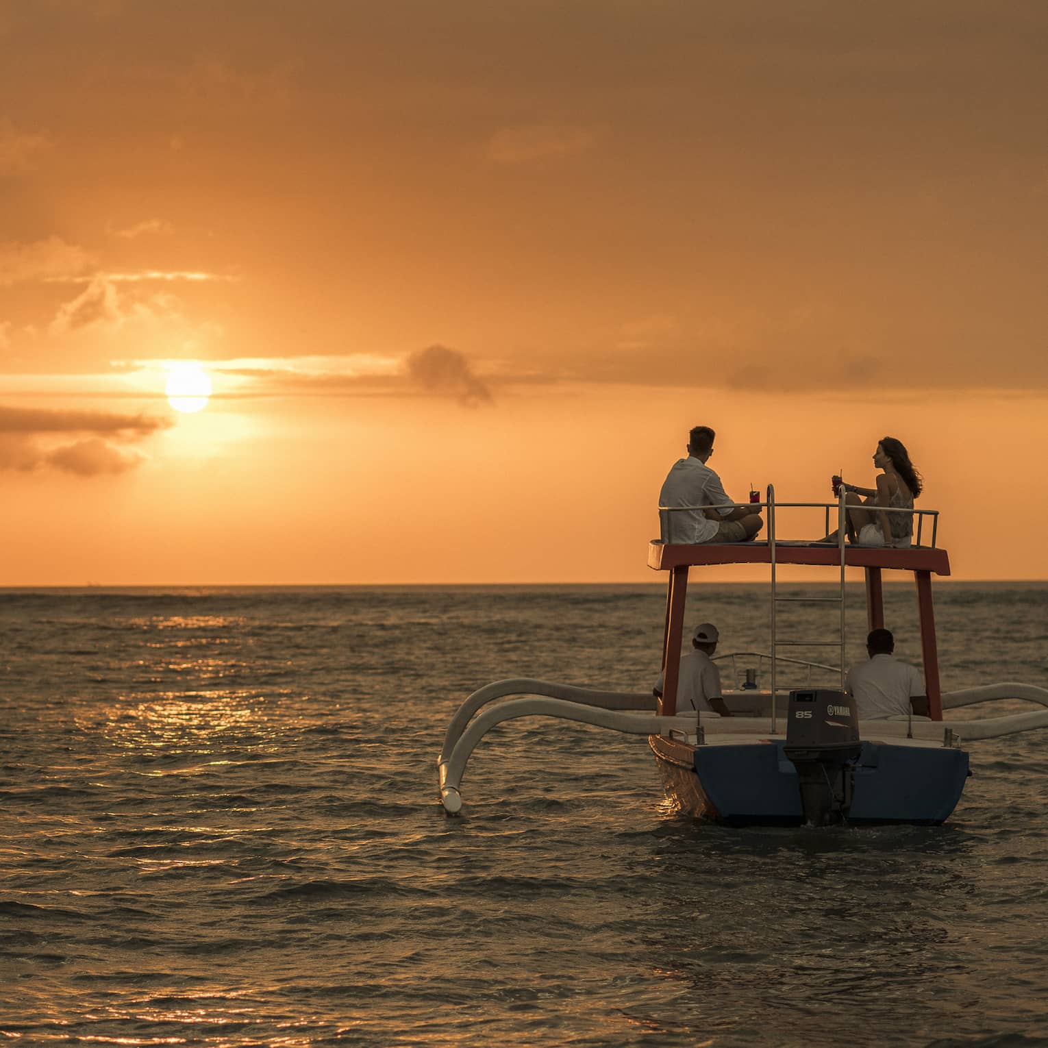 Couple sits on roof of boat sailing on ocean at sunset