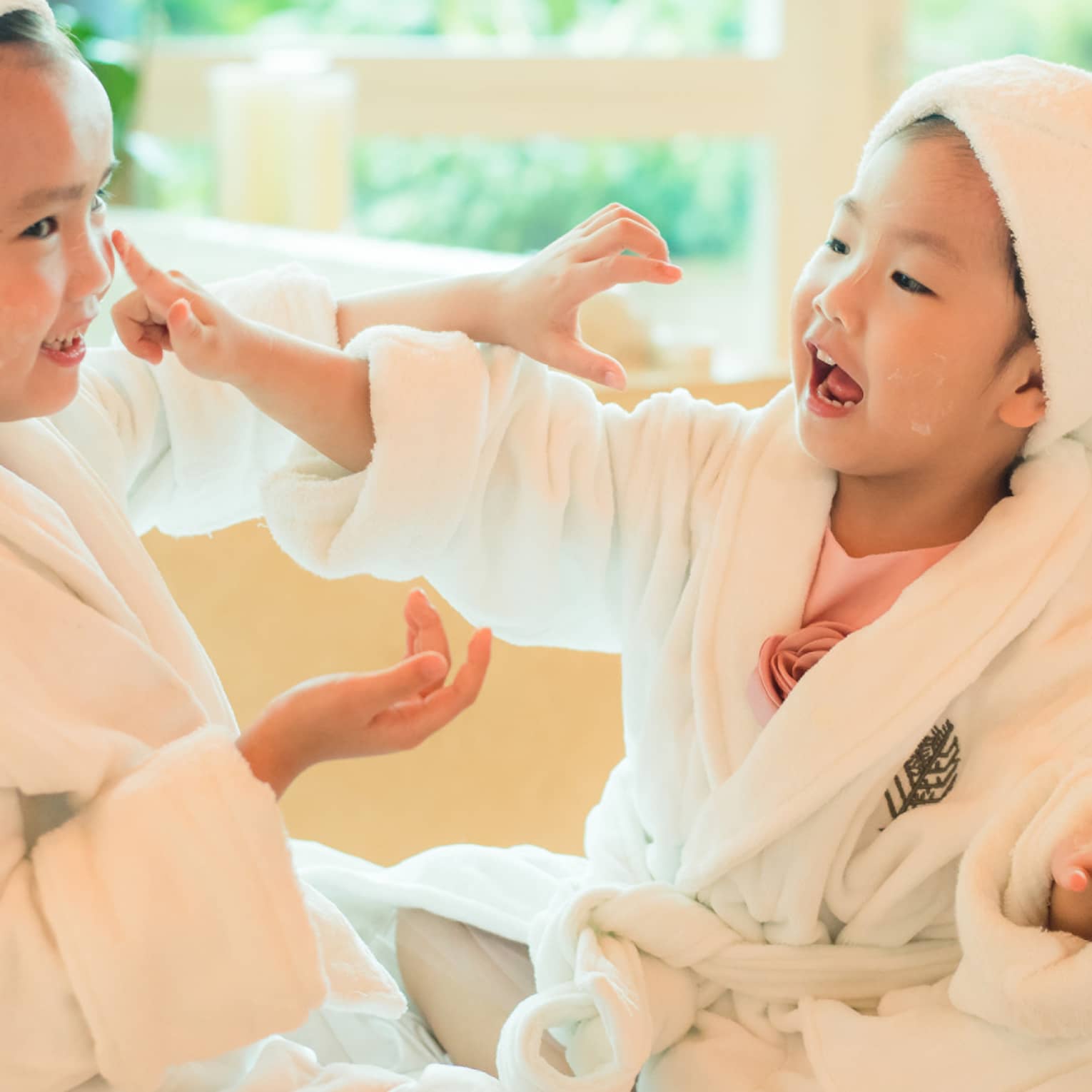 Two laughing young children in white bathrobes with towels around hair put lotion on each other's faces