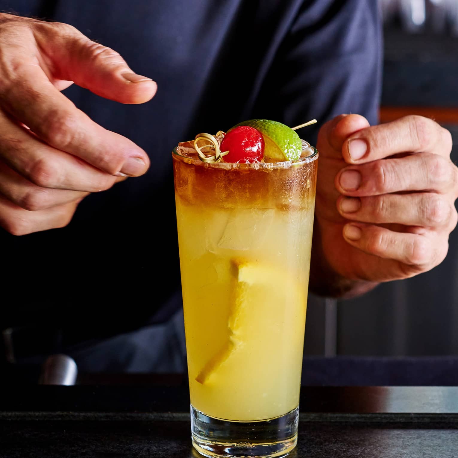 Close-up of hands adjusting a tall orange cocktail on ice, garnished with a cherry and a lime wedge on a small bamboo skewer.