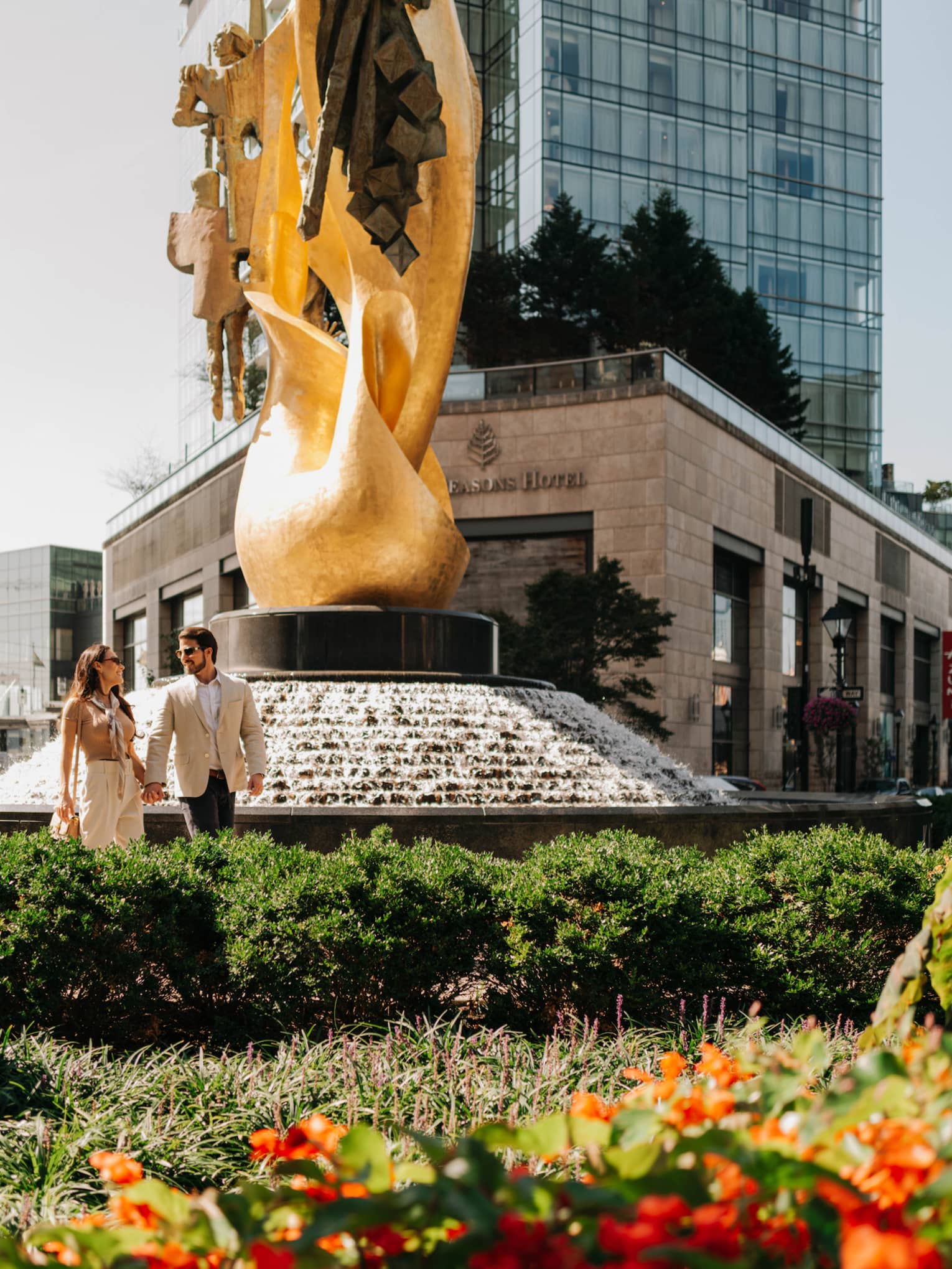 Man and woman walking in city near a statue and greenery.