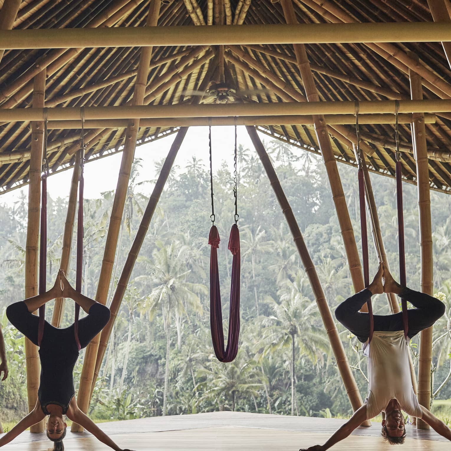 Woman and man hang upside-down on yoga hammocks under wood pavilion as instructor looks on