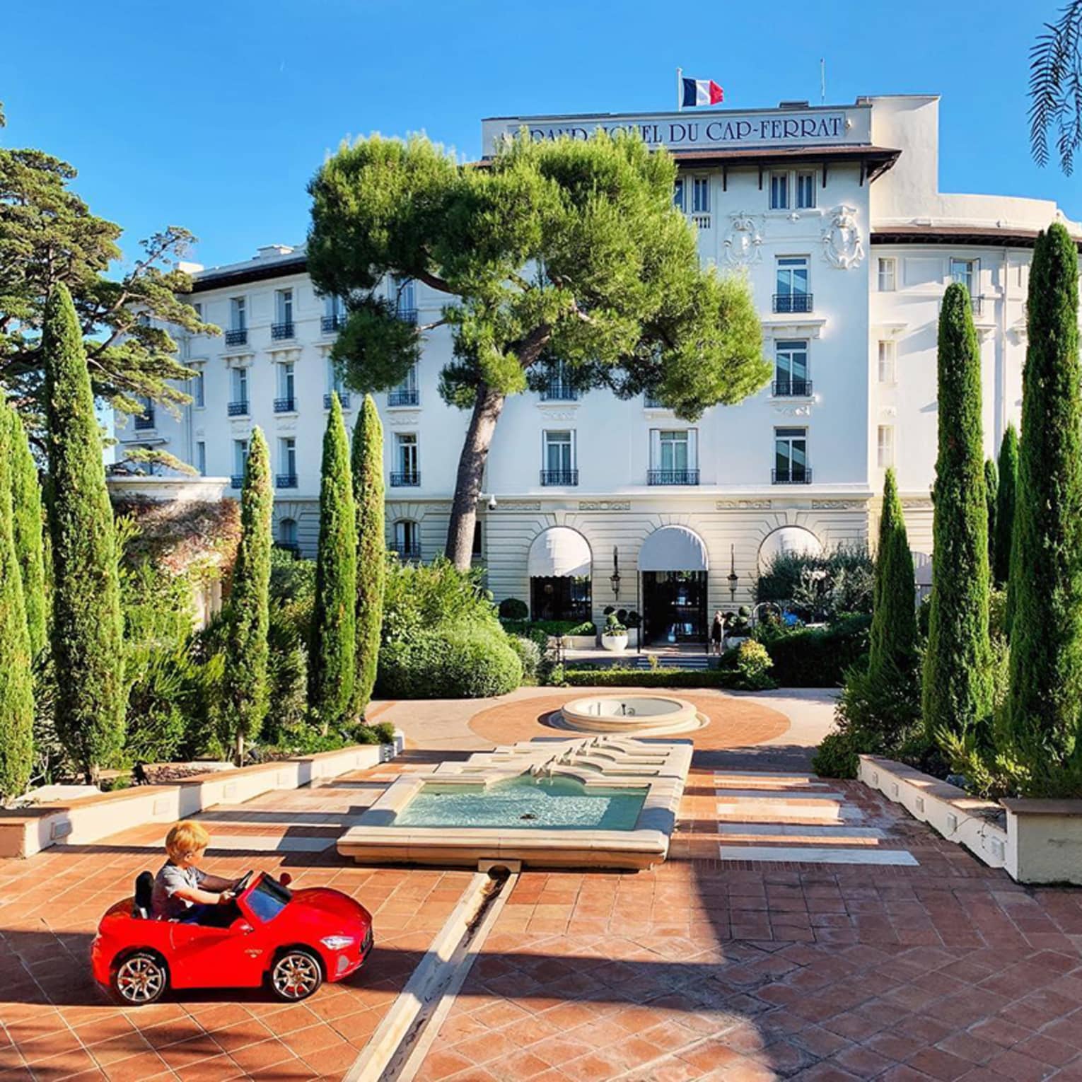 Young child rides in small red toy car in hotel courtyard with fountains, landscaped trees
