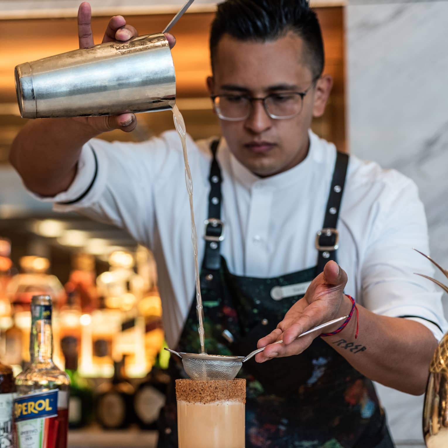 A man in a Four Seasons apron pours a drink through a sifter into a tall glass.