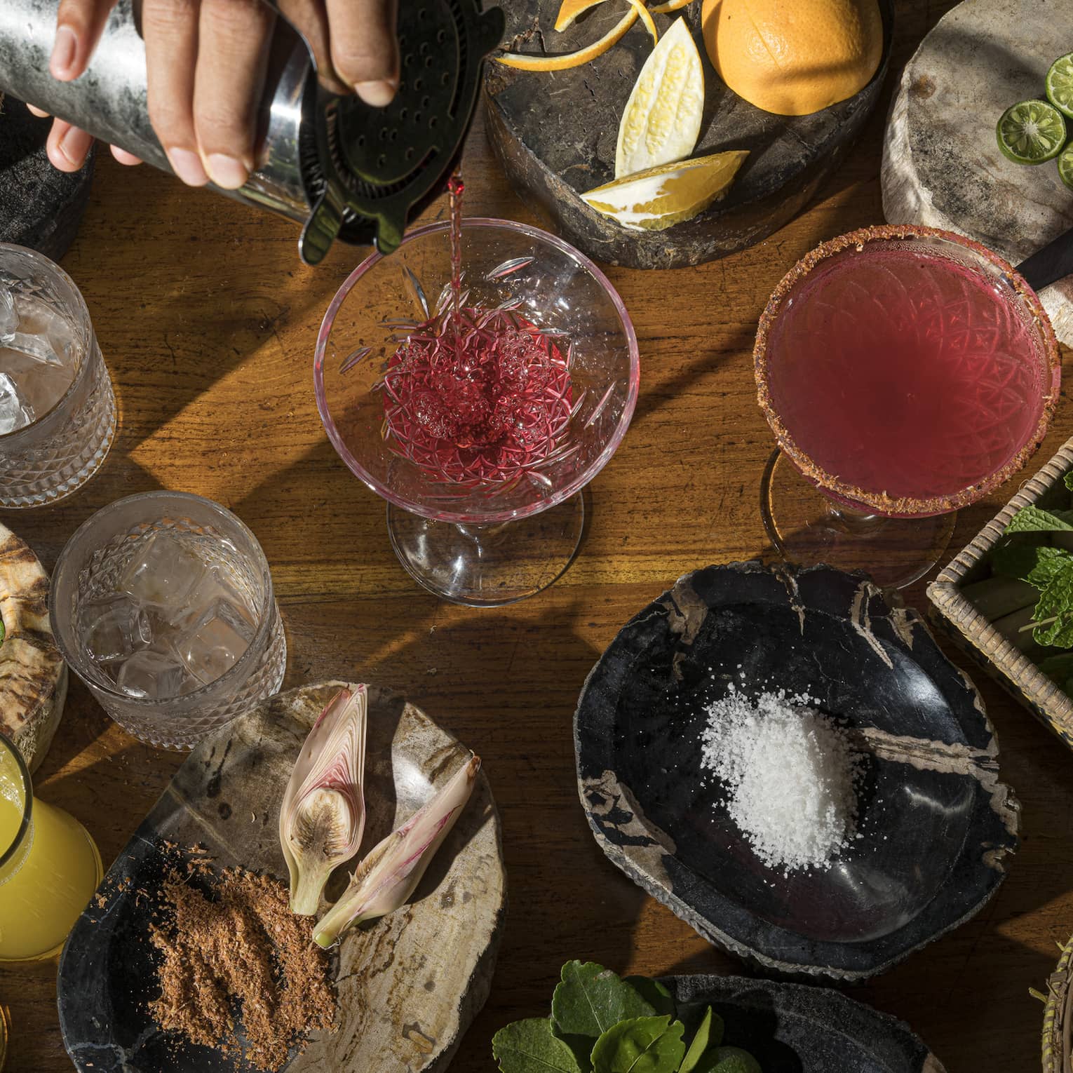 Aerial view of bartender's hand pouring ruby-coloured cocktail intro stemmed glass, local ingredients on wooden bartop surrounding it