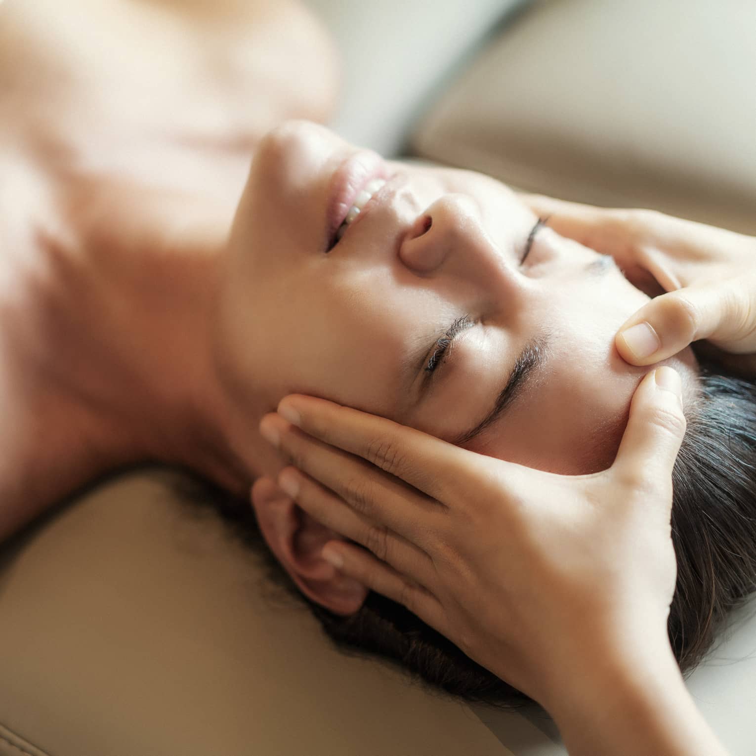 Spa facial, two hands rest on woman's forehead as she closes her eyes, lays under sheet on treatment table