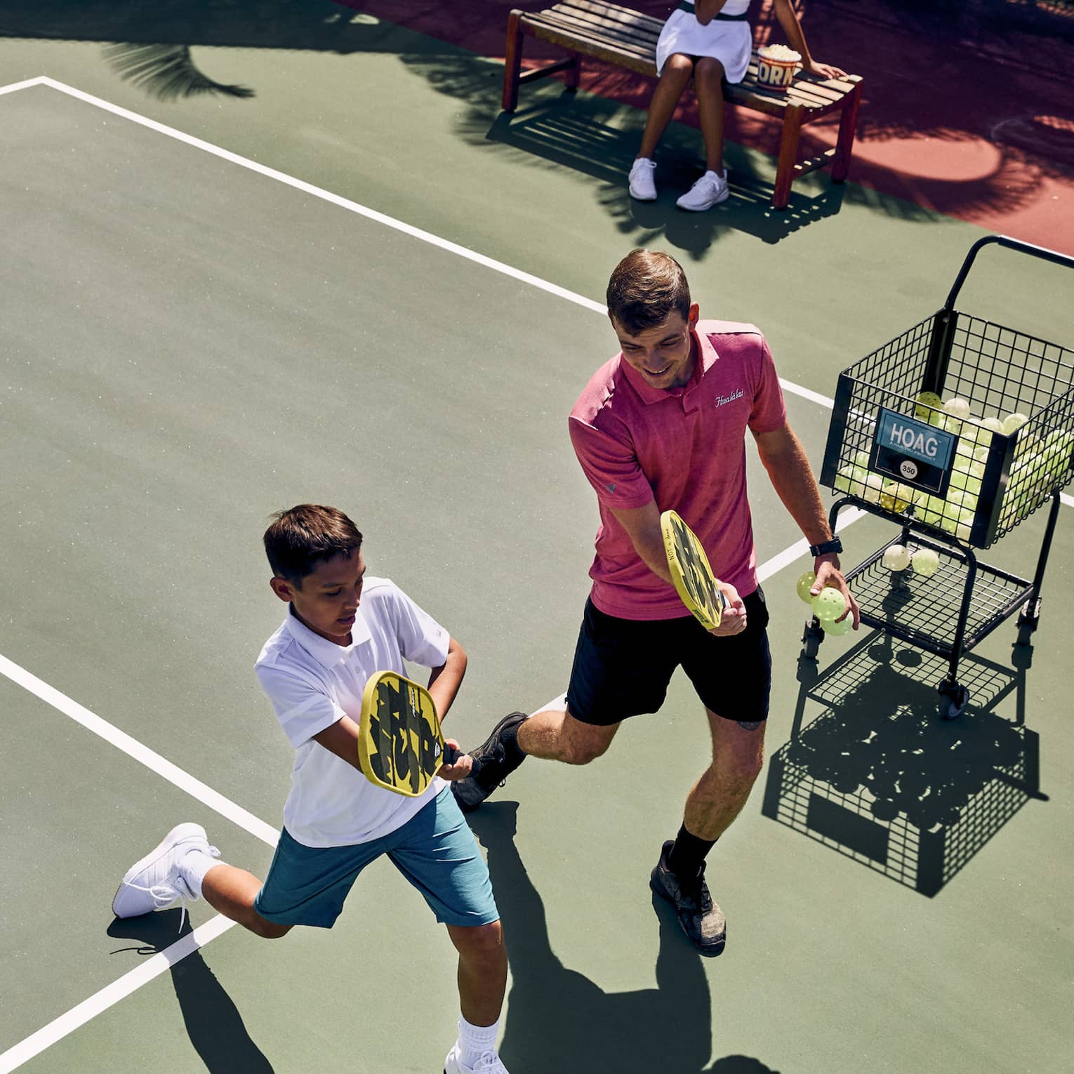 Aerial view of a pickleball pro instructing a player, both holding paddles while lunging forward, ball cart off to the side.