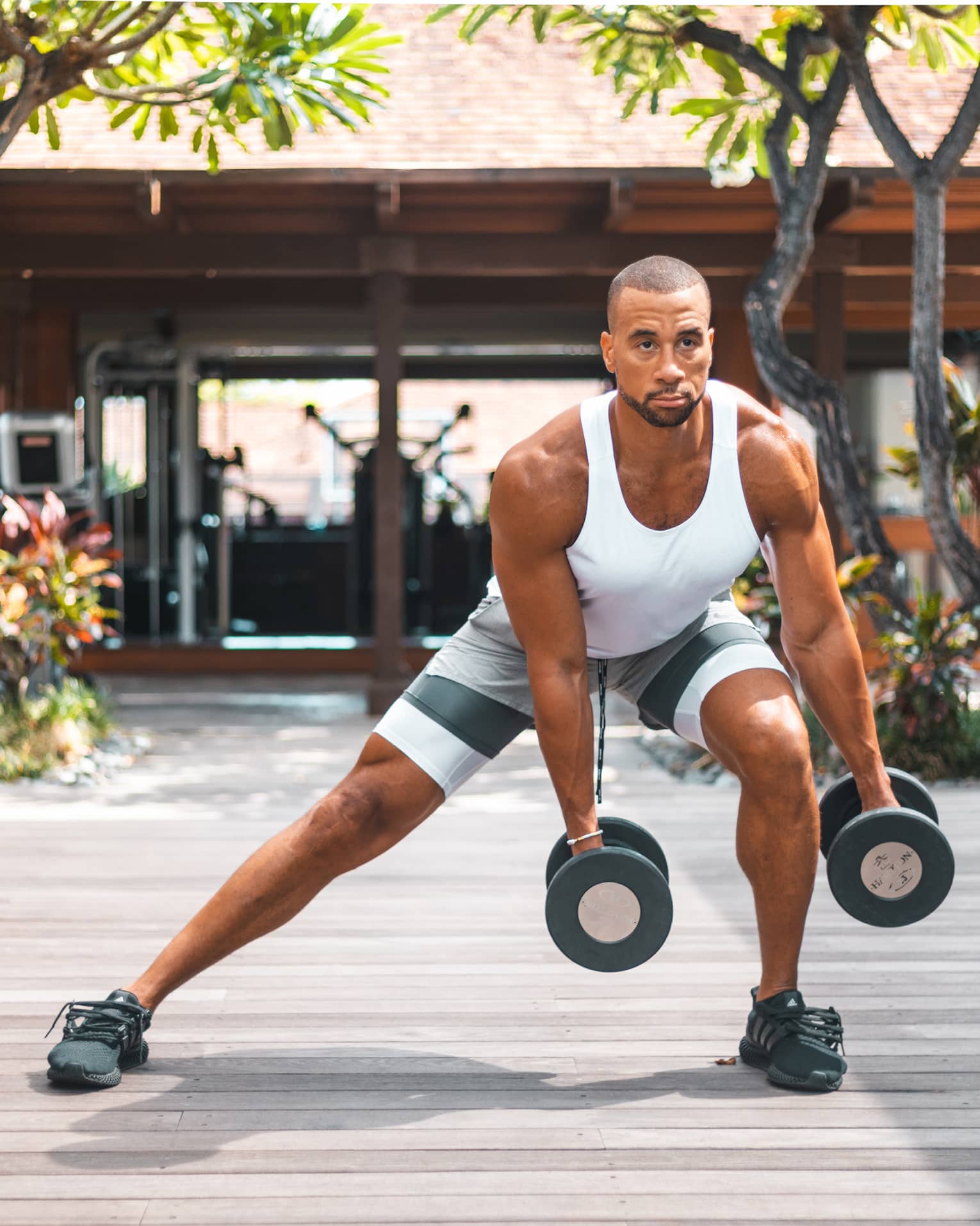 Man in white tank top and shorts doing side lunge on outdoor decking, holding weights