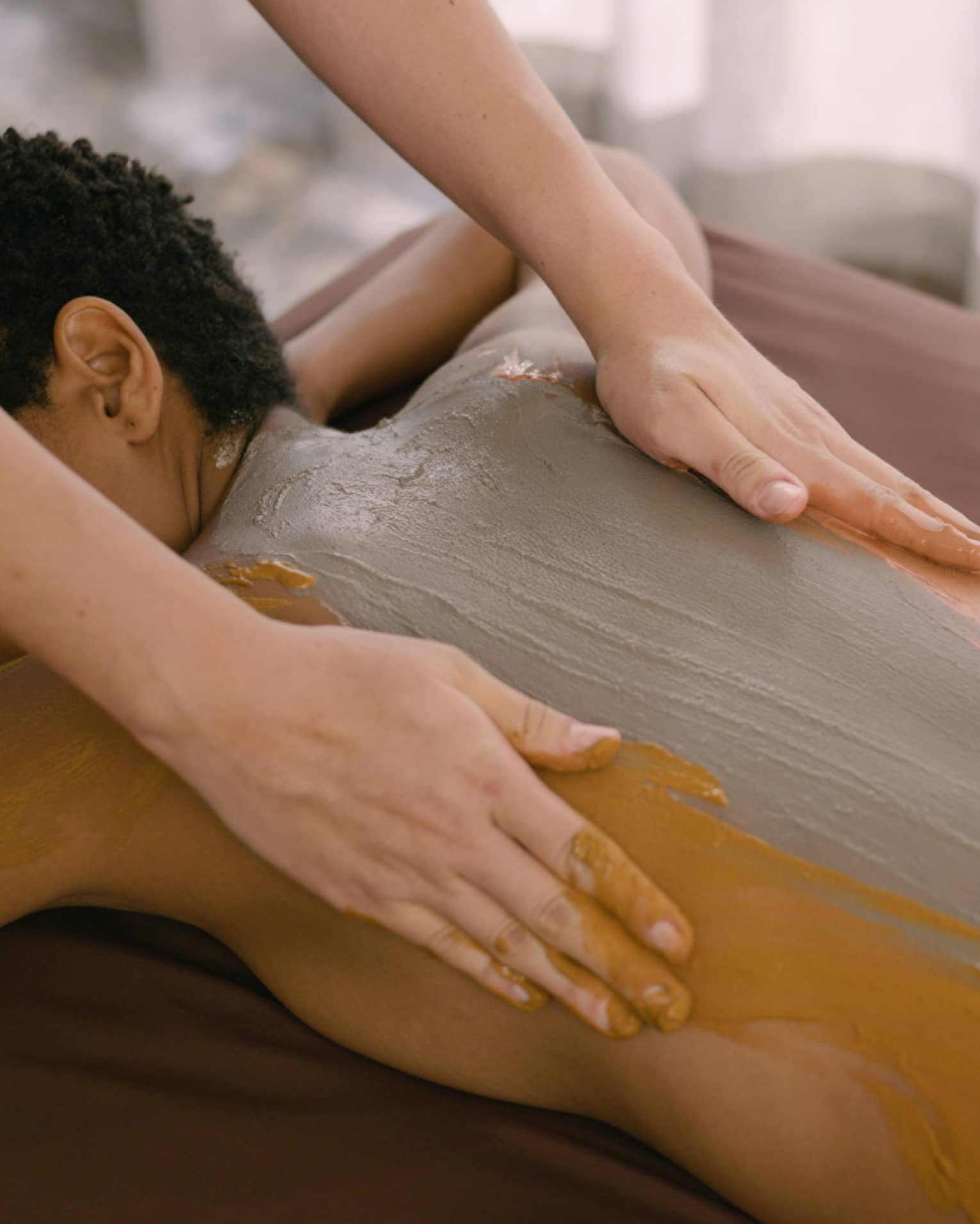 Hands rub clay across person's back as they lay on massage table
