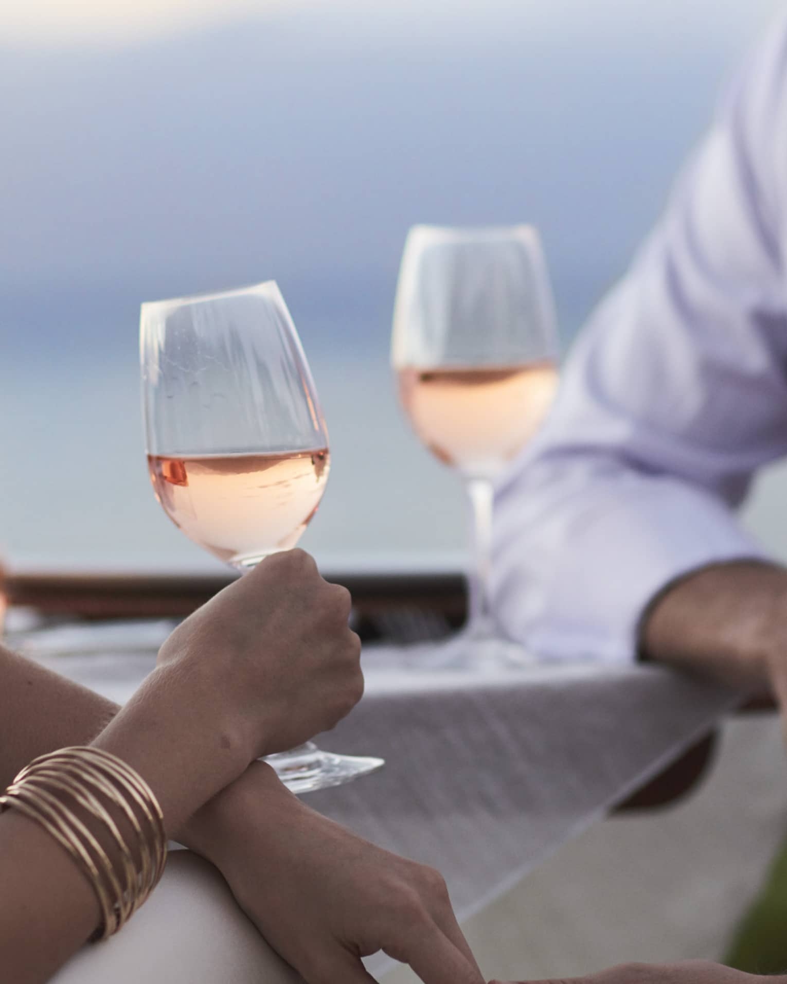 Close-up of woman and man in white, sitting and holding glasses of rose wine at dusk