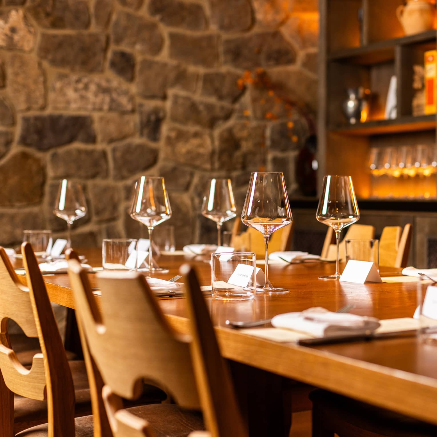 Table set for six in a dining room with a stone walls and a shelf filled with wine glasses