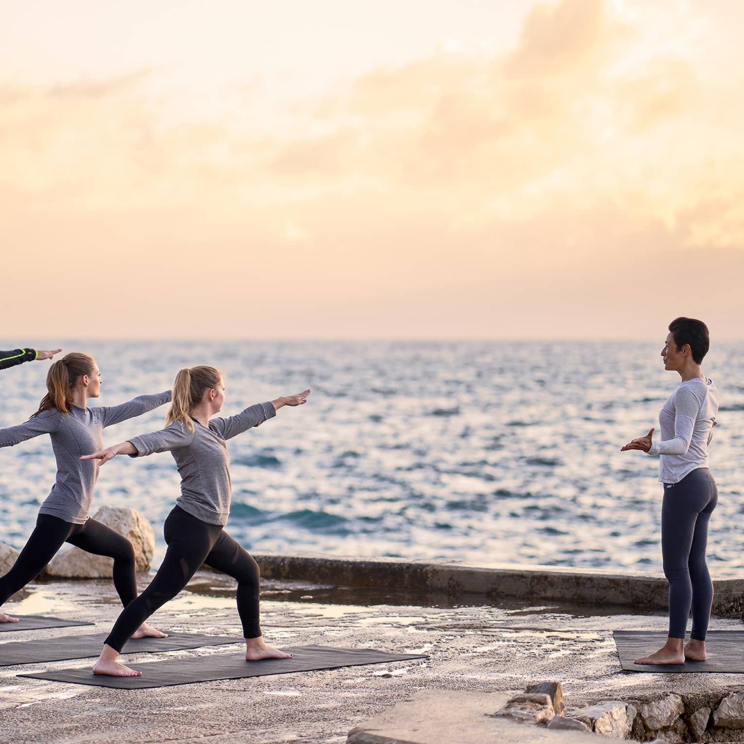 Three people in warrior yoga pose and coach standing at sea's edge at sunrise