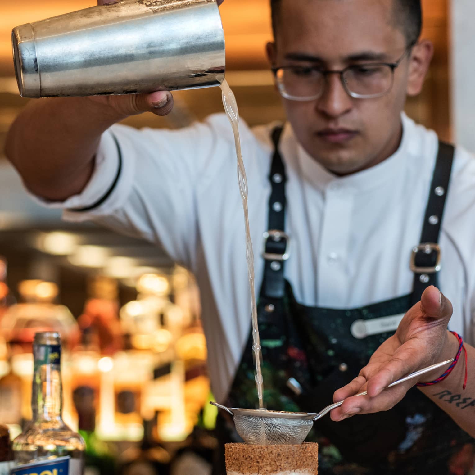 A man in a Four Seasons apron pours a drink through a sifter into a tall glass.
