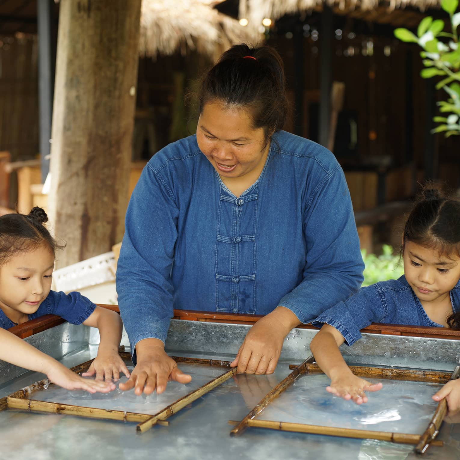 An adult stands between two children, running their hands along a clear, wet substance enclosed in two thin bamboo frames.