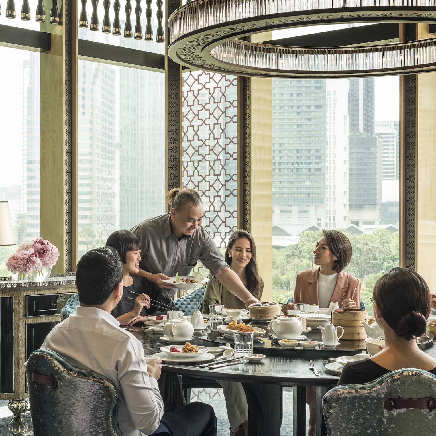 Seven diners sit at a table ensconced by glass walls as a server presents a dish in one hand, bamboo basket in the other.
