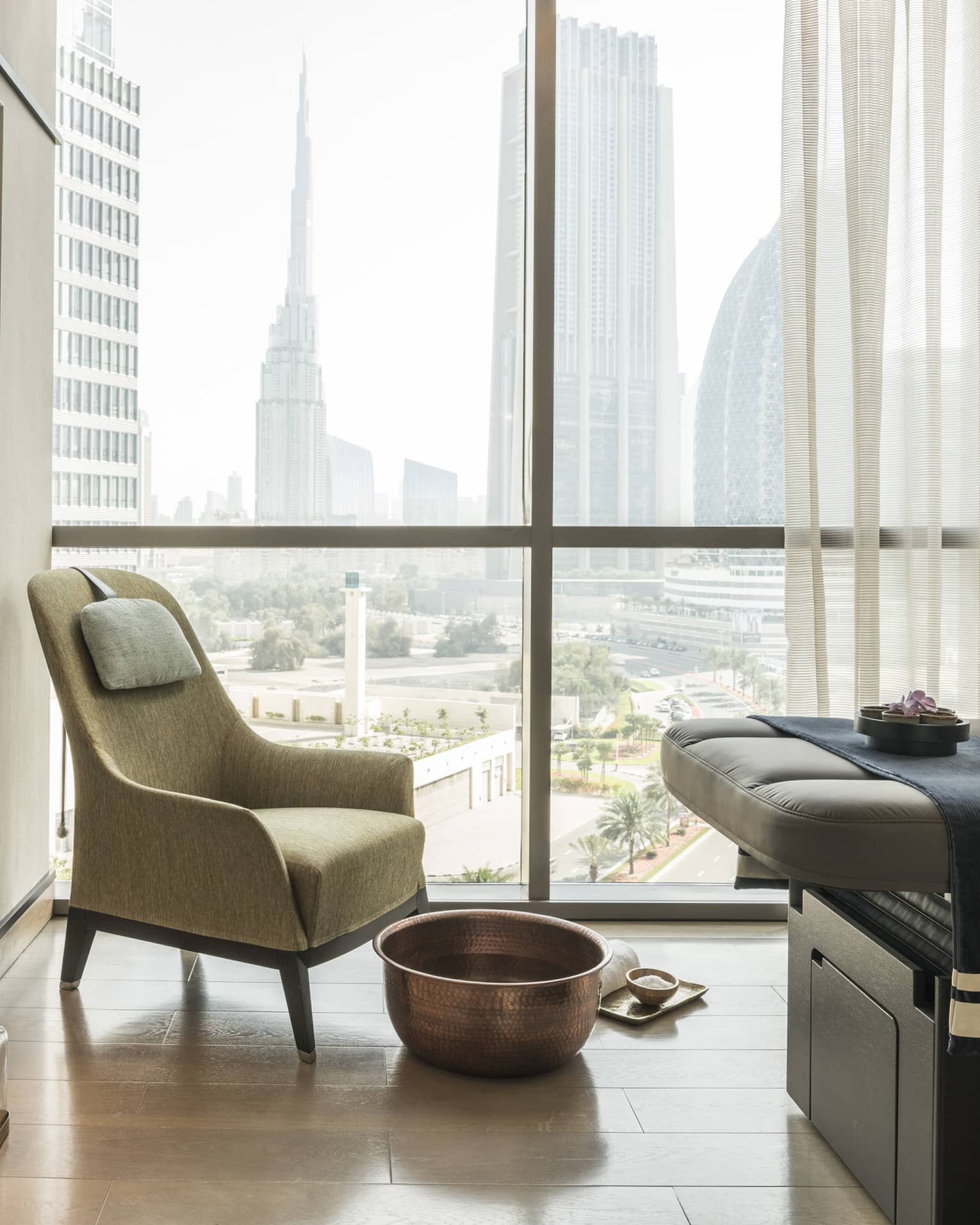 Armchair in hotel room corner by sunny window, large brass bowl foot bath, rolled white towel on floor