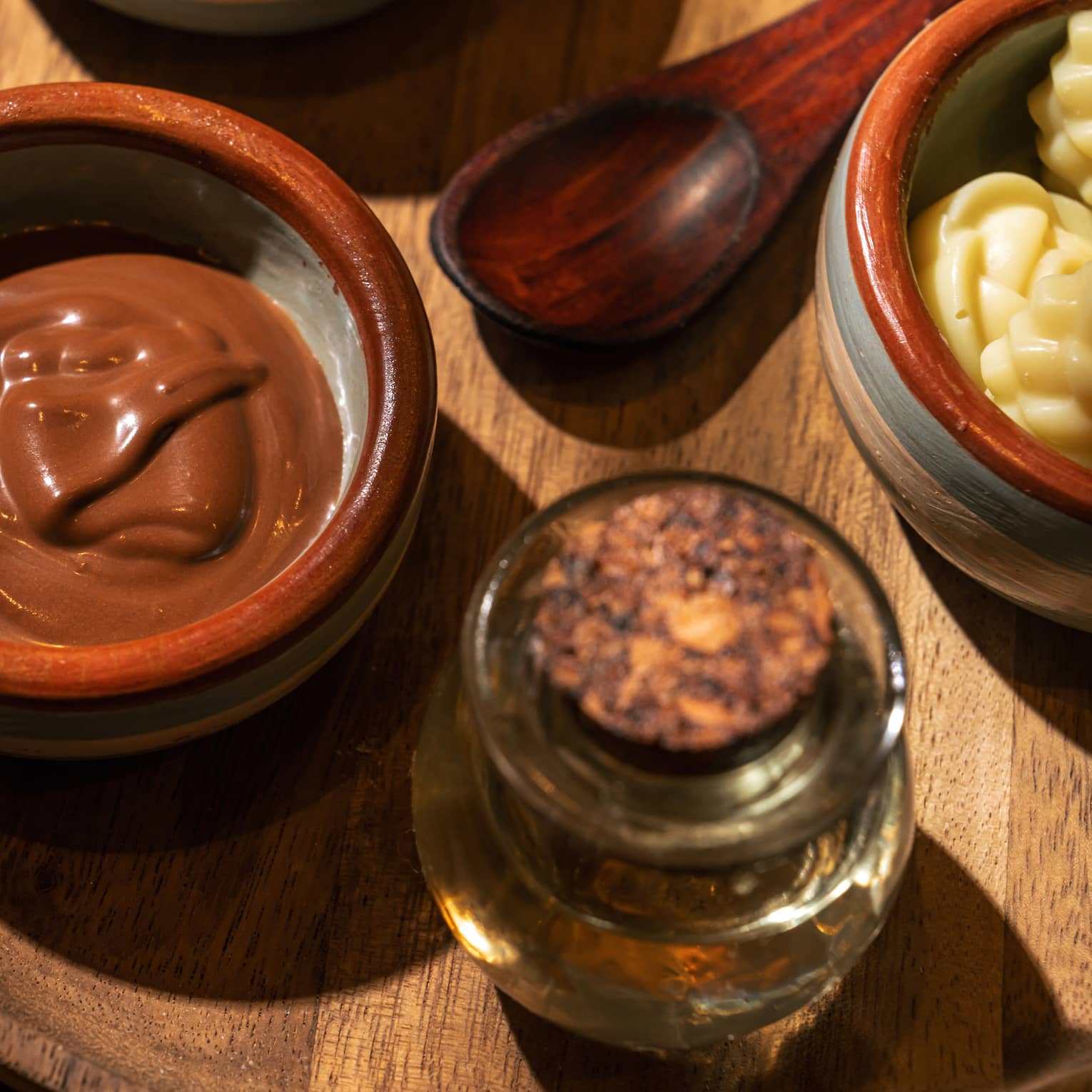 Stoneware bowl holding spa mud, wooden spoon, a glass bottle holding oils and another bowl holding soaps all sit on a round wooden platter