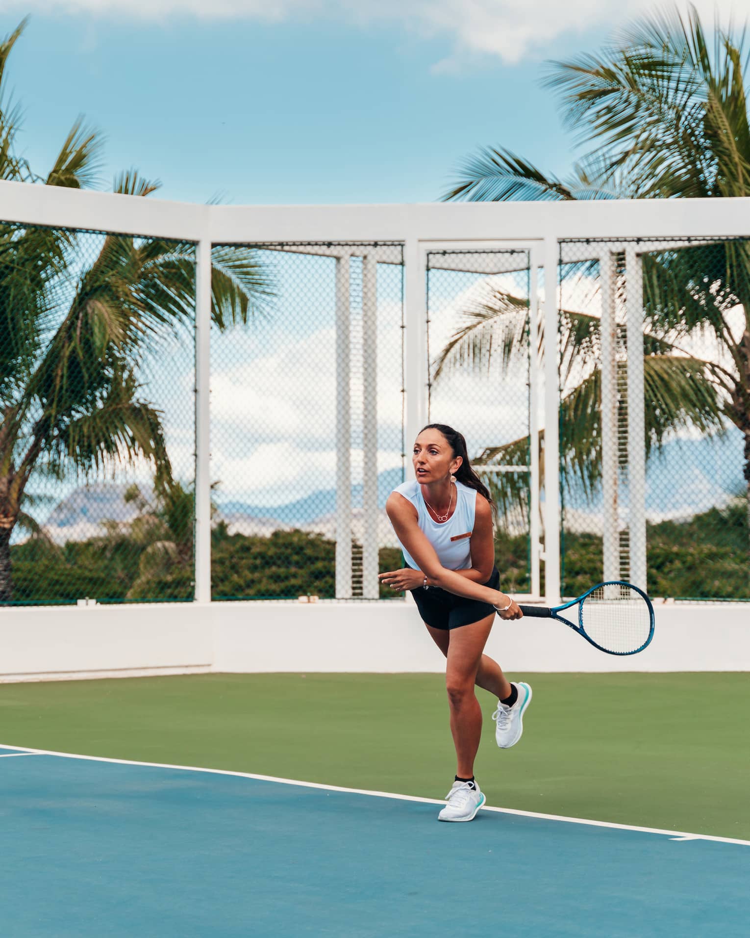 Front view of a player in follow-through after serving the ball on an outdoor tennis court, palm trees and mountains beyond.