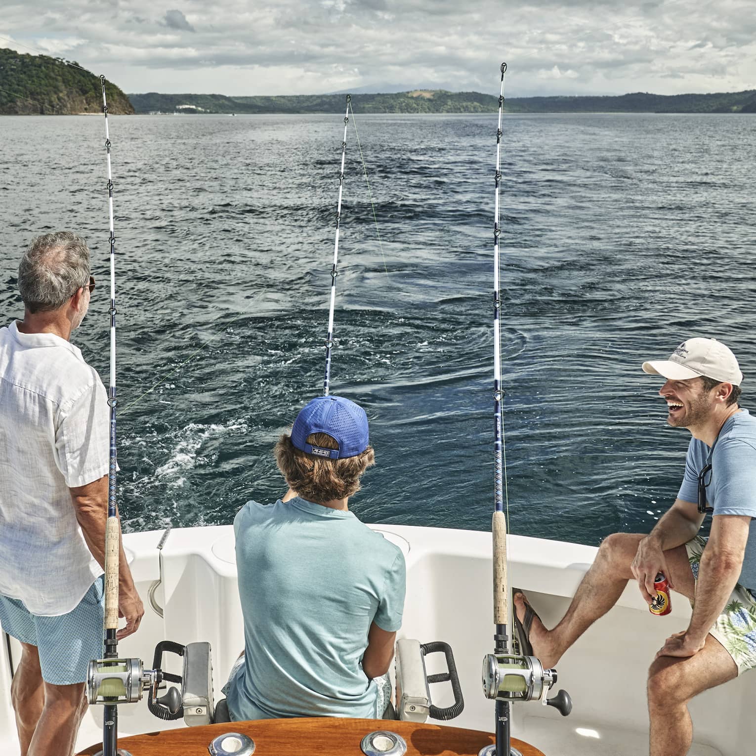 Three men of varying ages sit looking out at the water from the back of a boat outfitted with fishing rods
