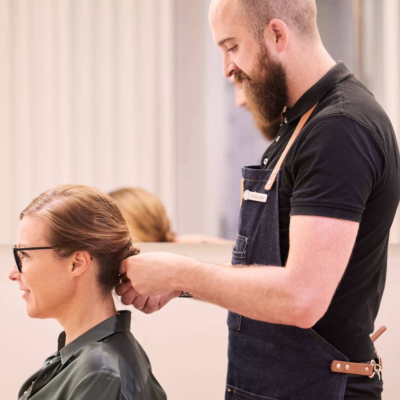 Woman sits in salon chair as hair stylist with long beard holds her hair