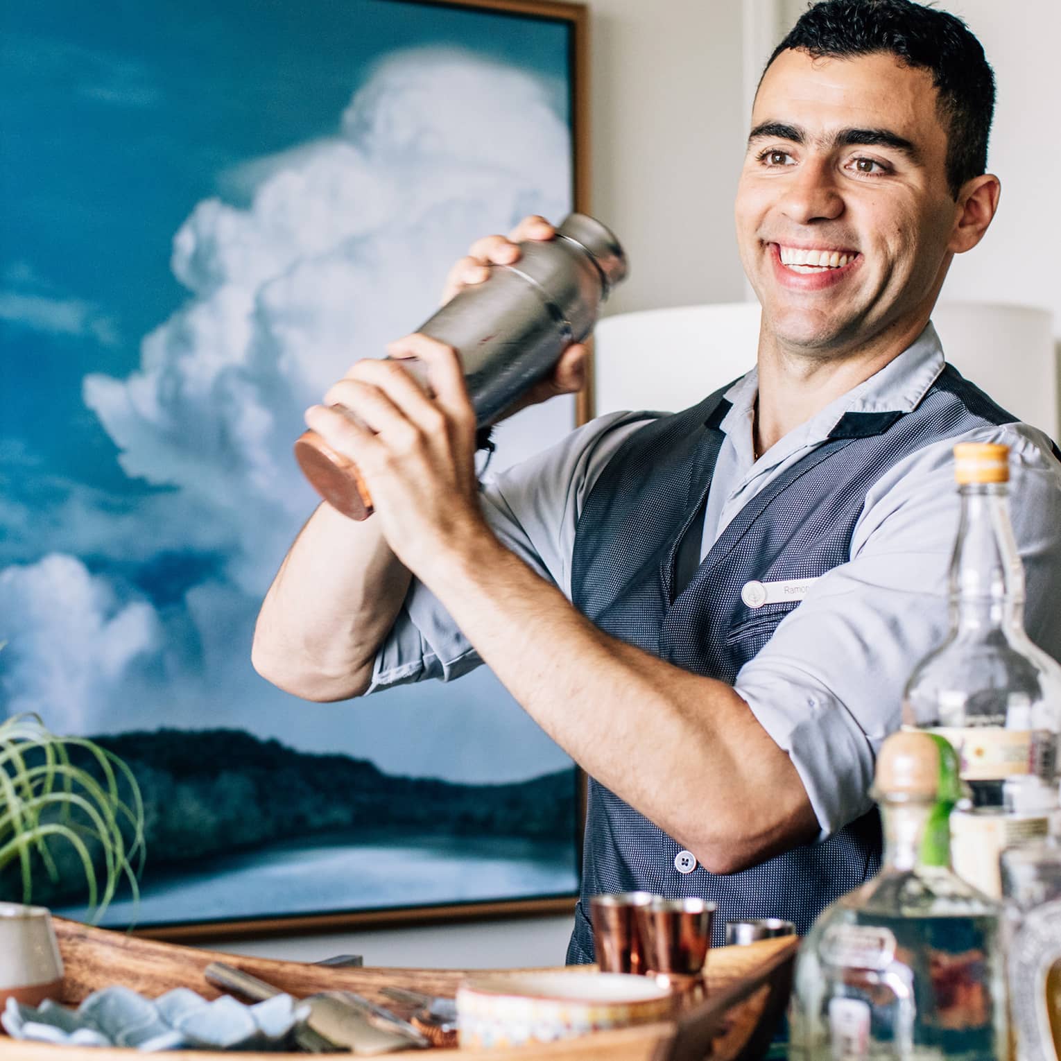 A bartender shaking a cocktail with liquor bottles on a cart in front of him.