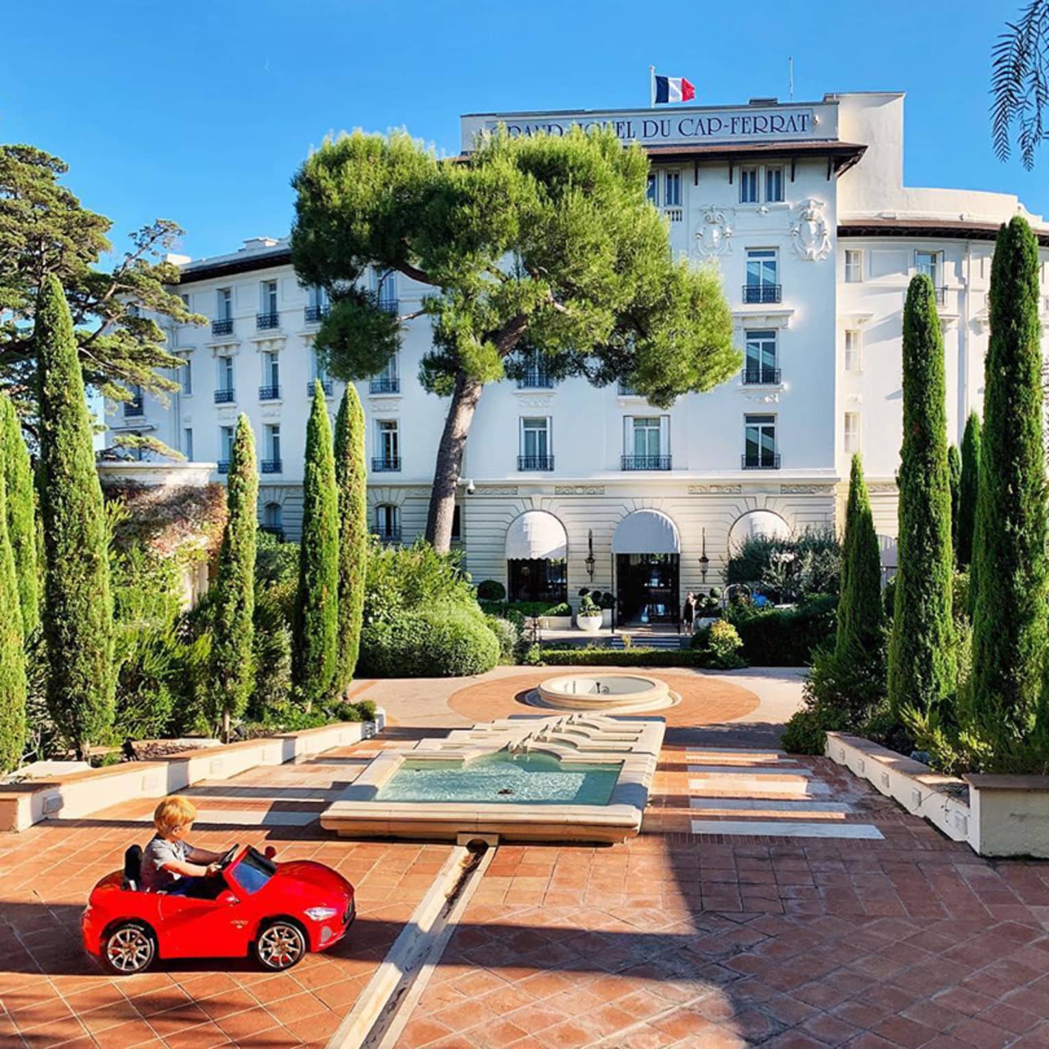 Young child rides in small red toy car in hotel courtyard with fountains, landscaped trees
