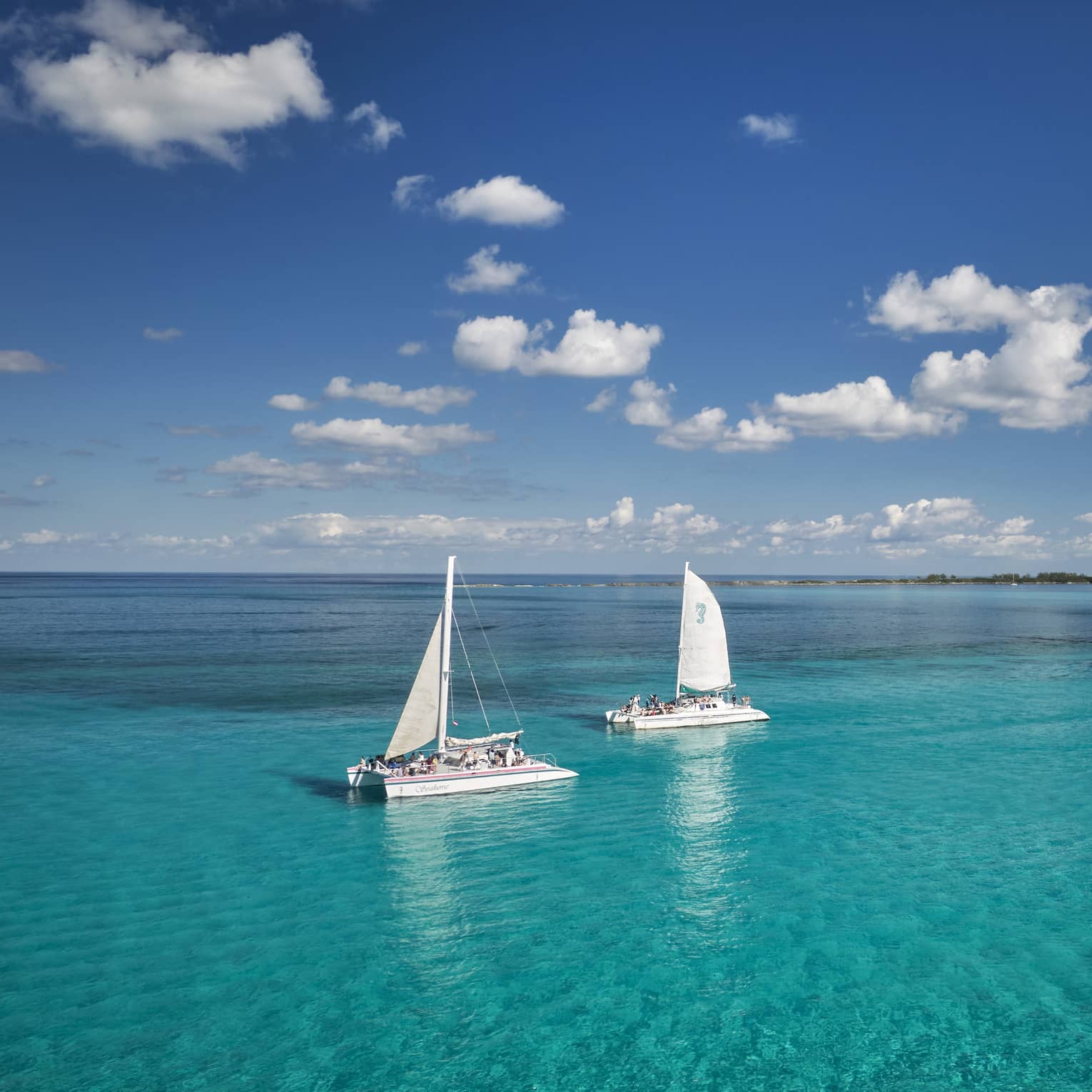 Guests aboard two catamaran sailboats glide through bright turqoise water, distant island beyond, under a cloud-dappled sky.