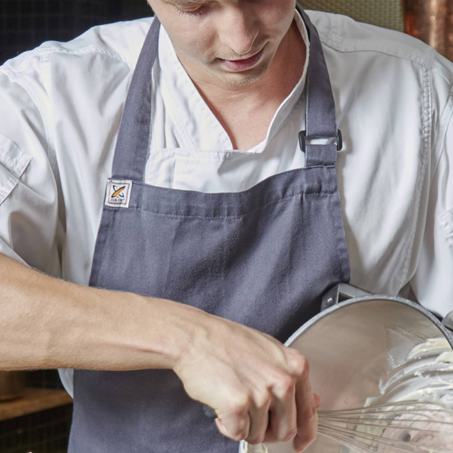 An aproned chef whisks cake batter in a bowl over a pebbled countertop, by a rolling pin and jar of white chocolate pieces.