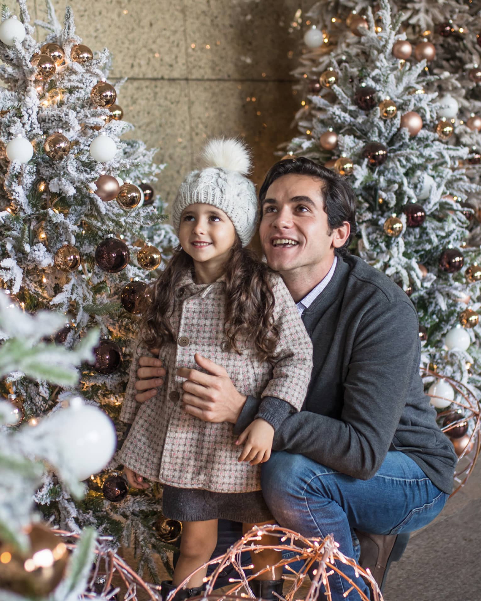 Man crouched with arms around girl in coat and hat admiring Christmas tree