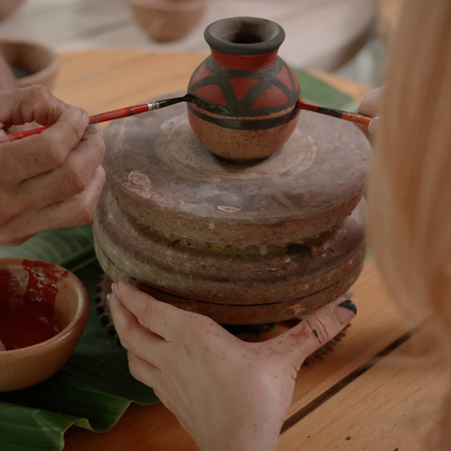 Two sets of hands using thin paintbrushes to decorate a small ceramic vase on a pottery wheel amid pots of paint and pigment.