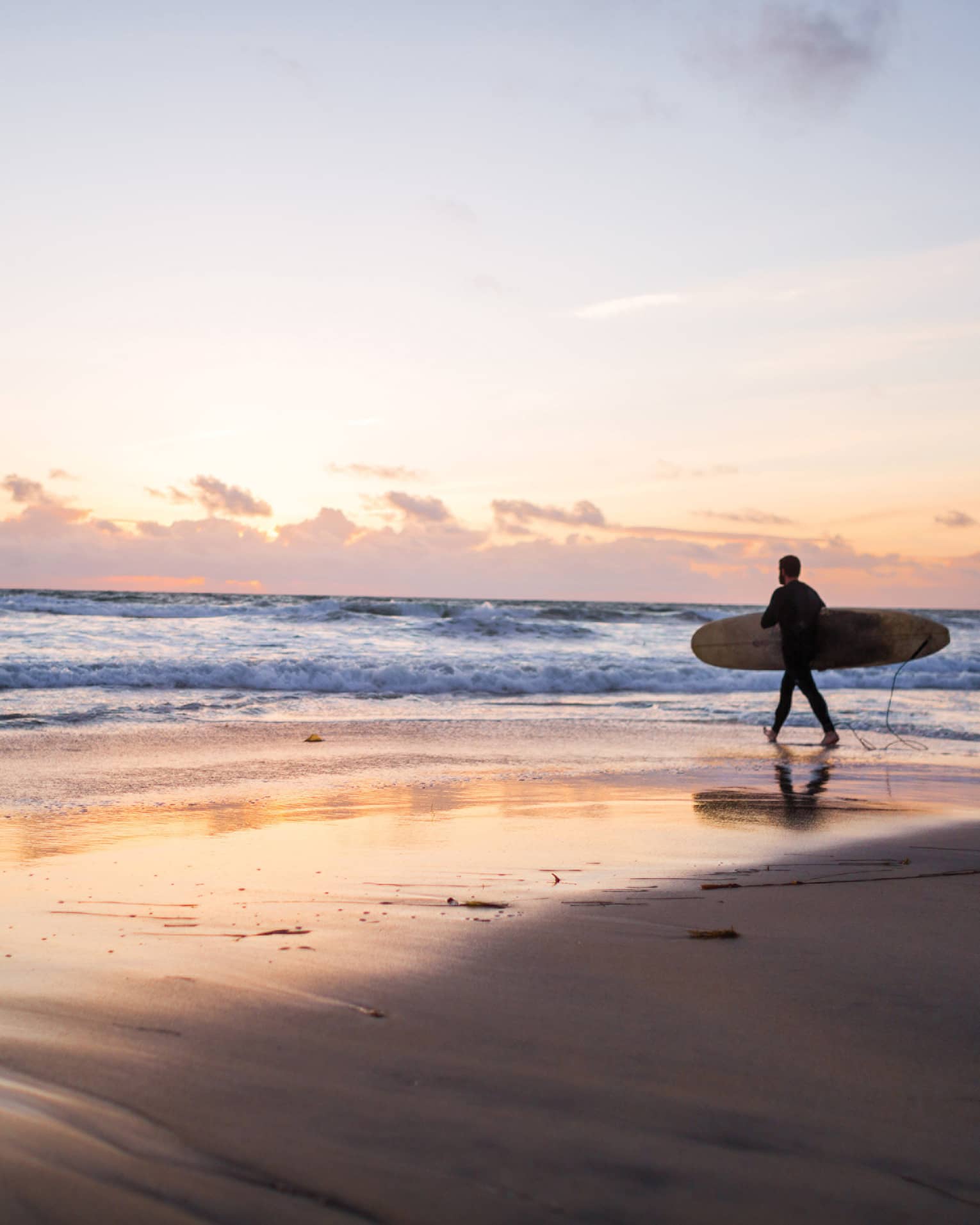 Under an expansive sky at sunset, clouds receding on the horizon, a surfer with a board heads into the glimmering ocean.