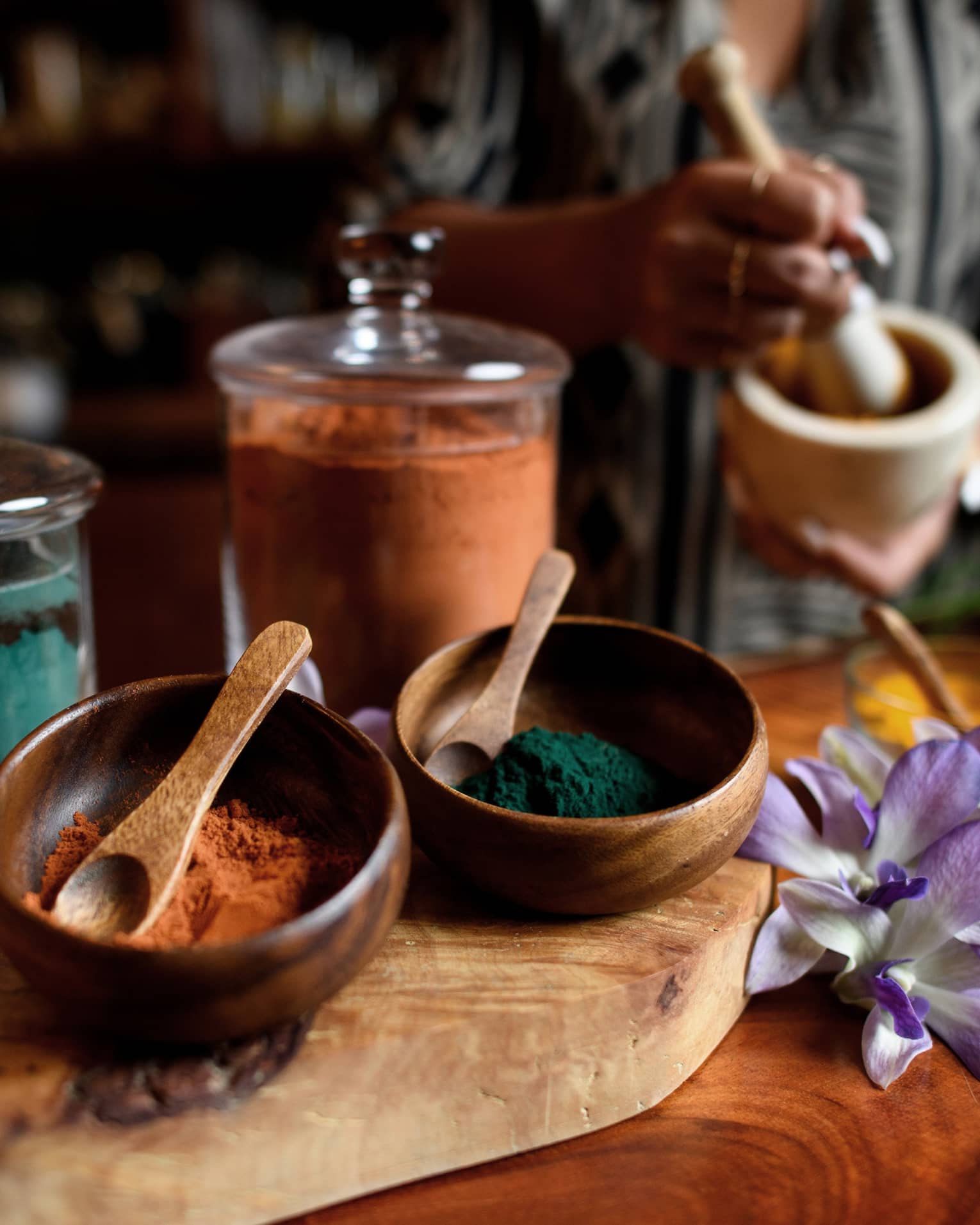 Small wood bowls and spoons beside glass jars filled with colourful powders, tropical flower