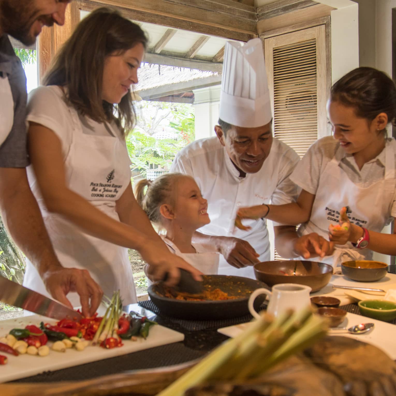 A smiling chef oversees a family joyfully indulging in a private cooking class at Four Seasons